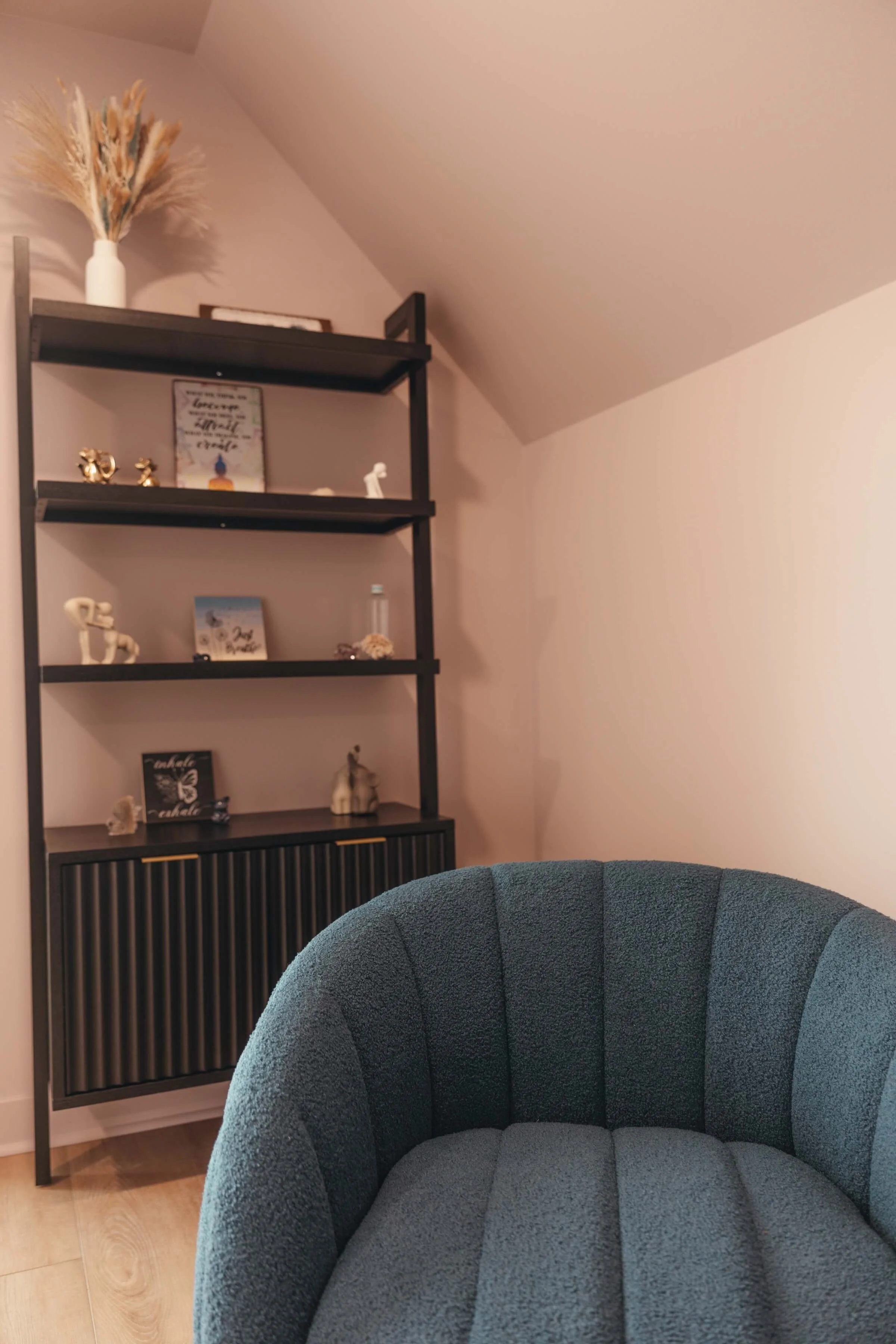Close-up of a teal upholstered armchair in a cozy living space with a black shelving unit holding decorative items, framed pictures, and books, against a beige wall with a sloped ceiling.
