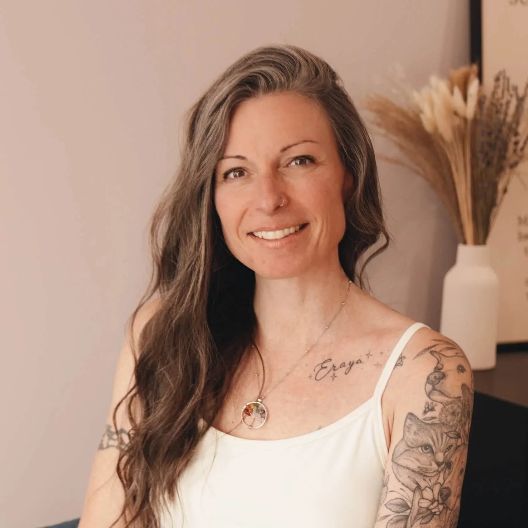 Portrait of a smiling woman with long brown hair, tattoos on her arm and chest, wearing a white tank top, standing indoors near a beige wall and a vase with dried flowers.
