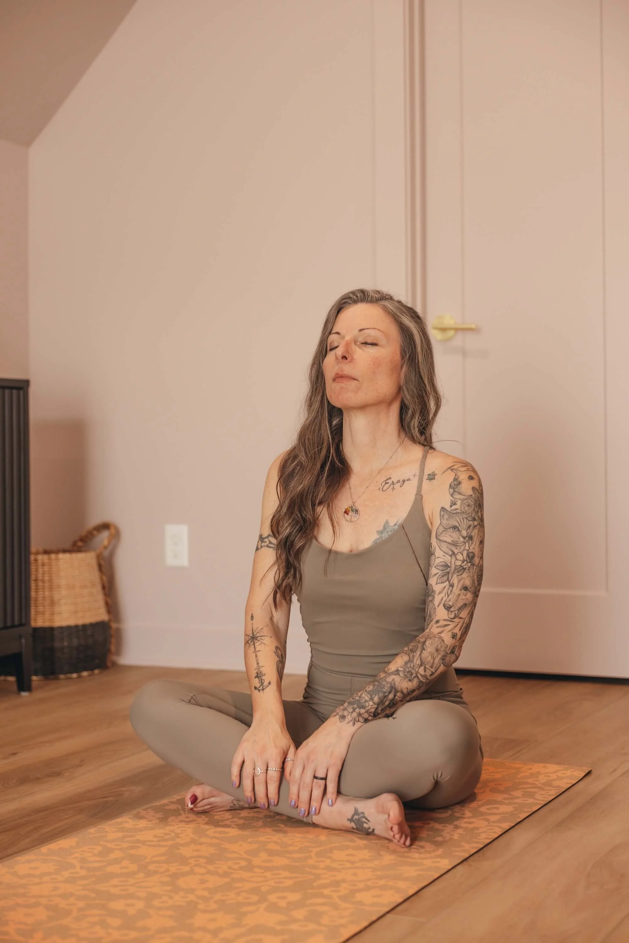 A woman with long gray hair and tattoos is sitting cross-legged on an orange patterned yoga mat in a room with hardwood floors and cream-colored walls, eyes closed, practicing yoga or meditation.