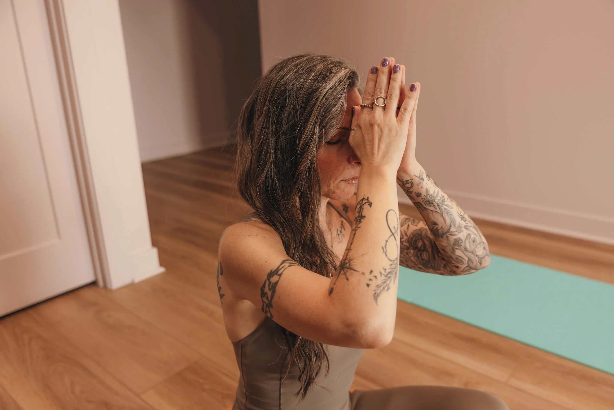 A woman with long gray and brown hair, covered in tattoos, sitting on the floor in a room with wooden flooring, practicing yoga or meditation with her hands in prayer position in front of her face. There is a light green yoga mat behind her.