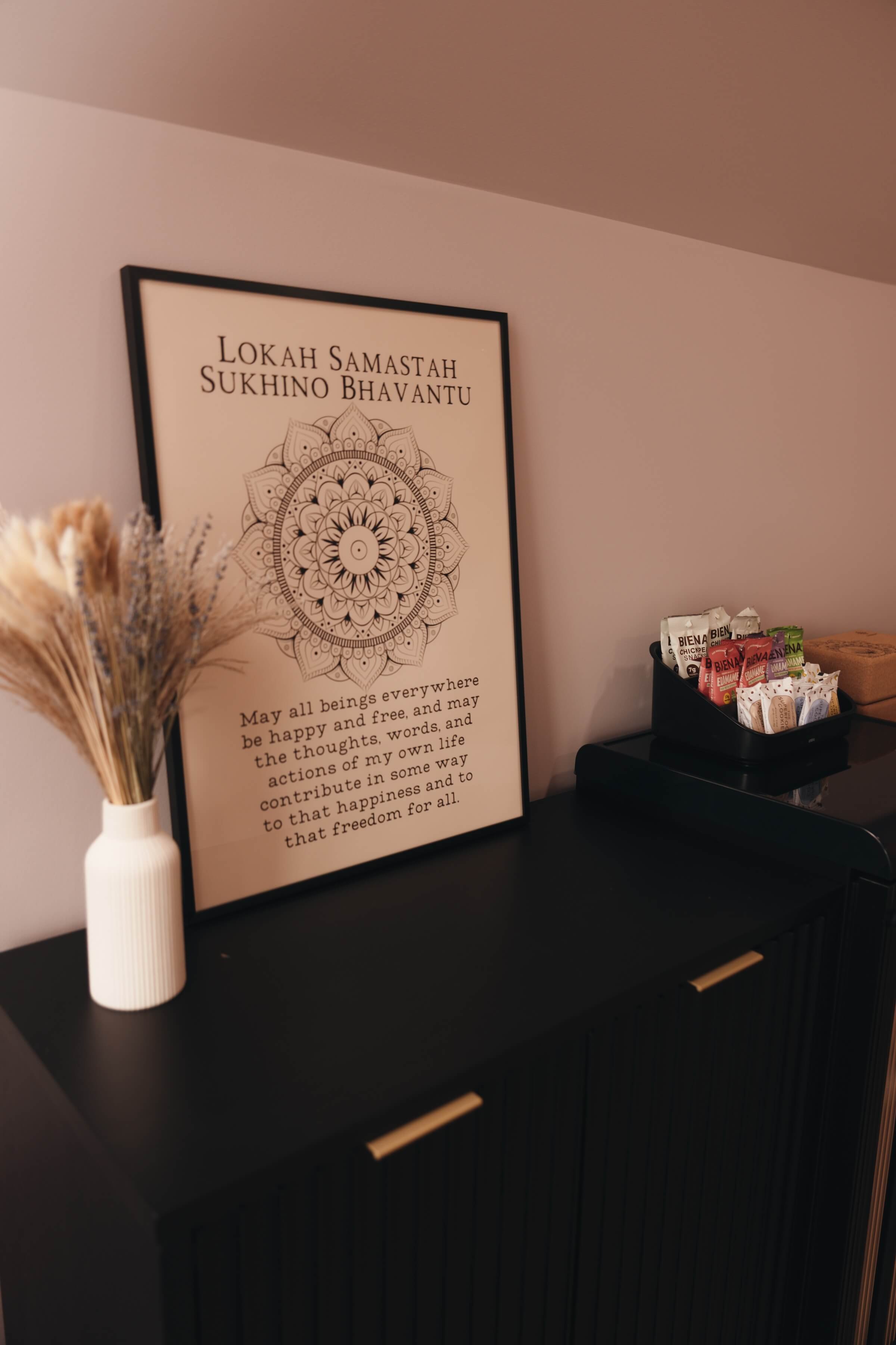 A framed quote on a wall that reads 'Lokah Samastah Sukhino Bhavantu', with a decorative mandala design on it, placed beside a white vase with dried flowers and a black container filled with snack packets on a dark wooden side table.