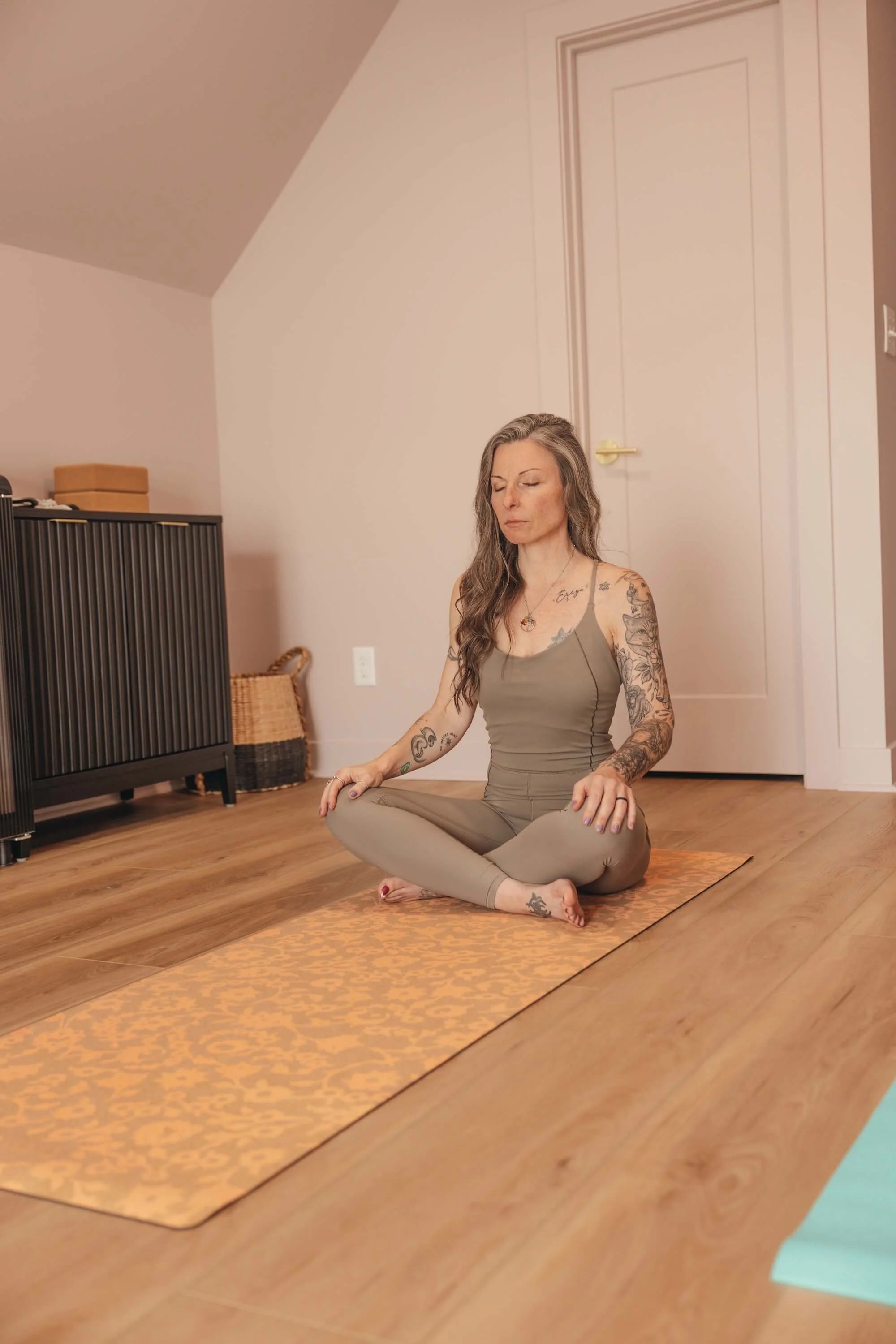 A woman with tattoos practicing meditation on an orange patterned yoga mat in a room with wooden floors and beige walls.