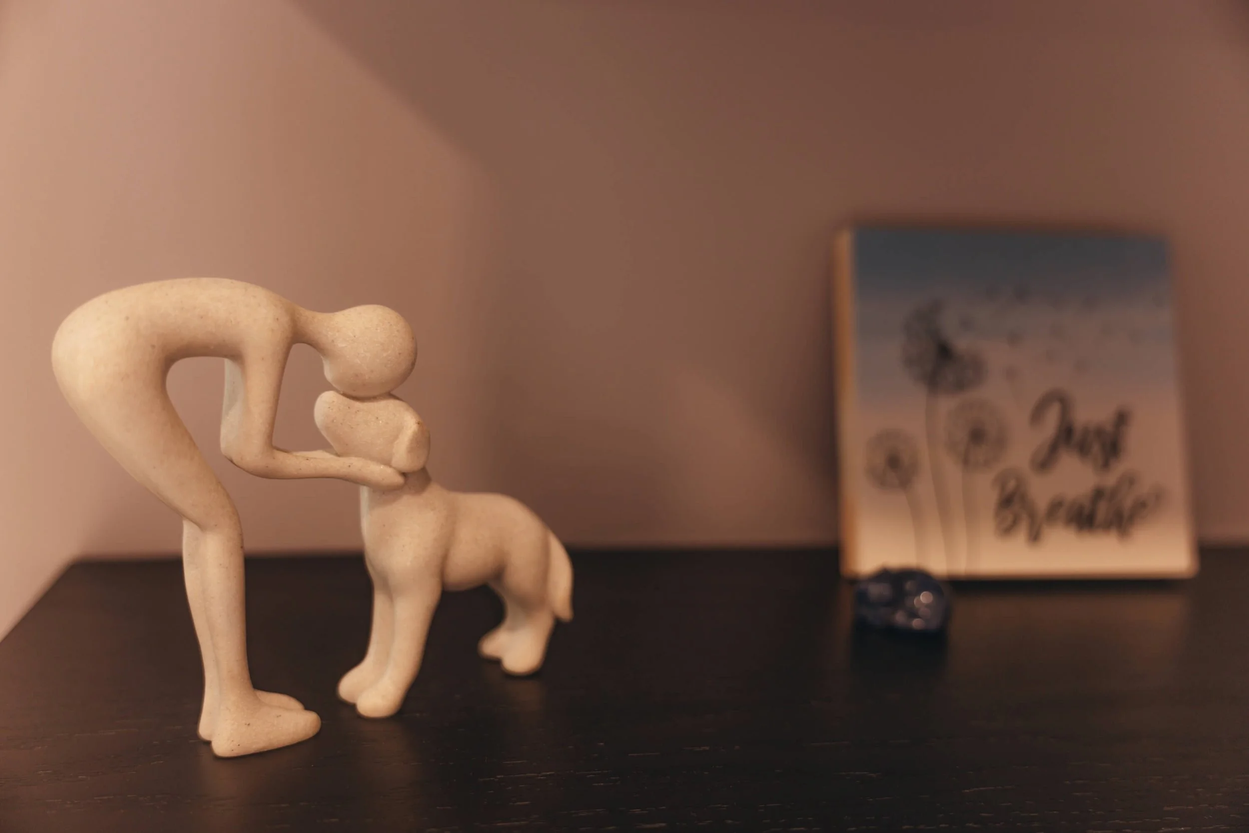 Decorative figurines on a black surface with a blurry background, including a beige elephant and a person figurine leaning on it.