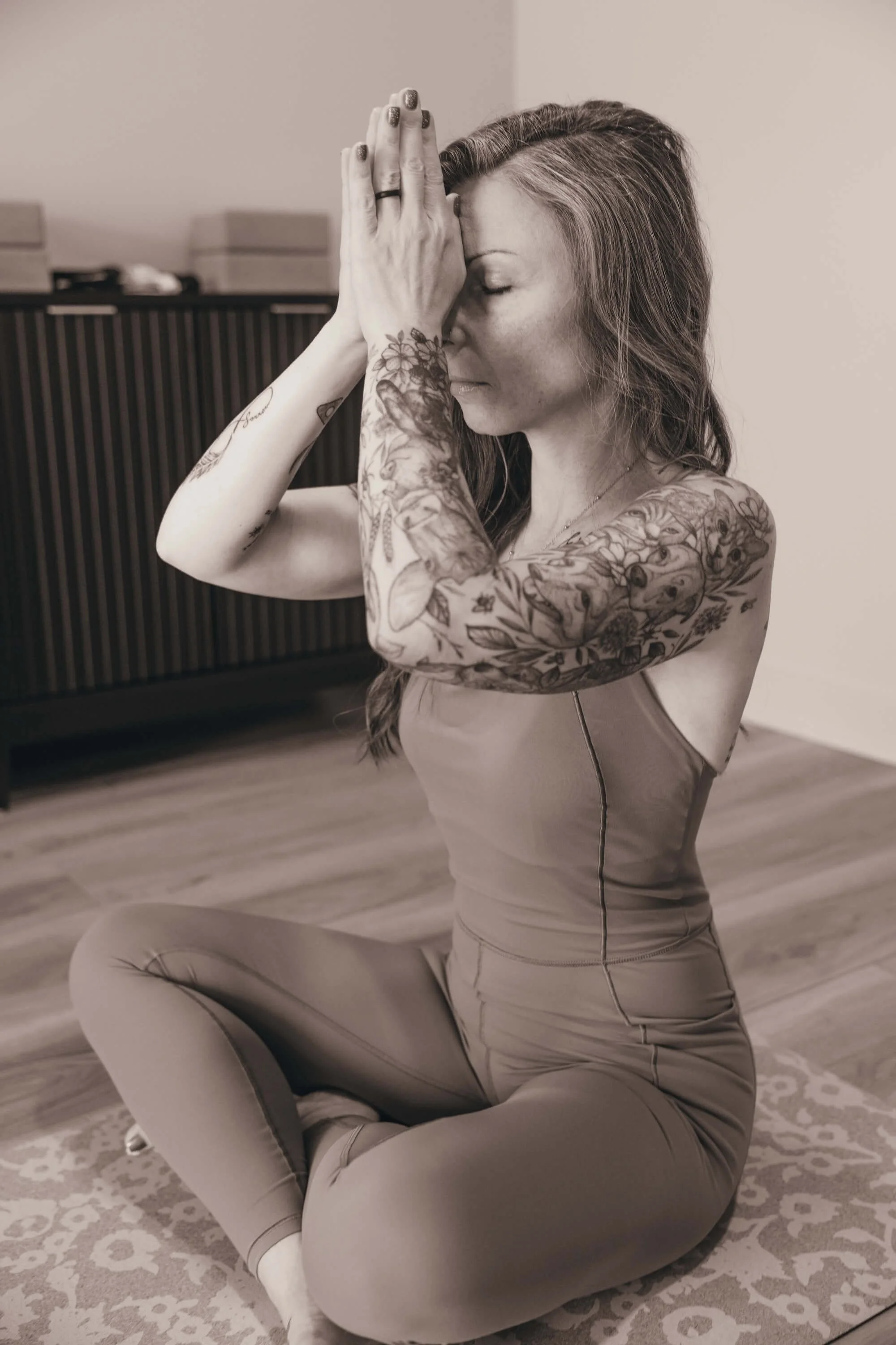 A woman with tattoos on her arms and long hair in a bun is practicing yoga indoors, sitting on a decorative rug with her right knee on the floor, her left foot flat on the floor, and her hands pressed together in prayer position against her face with her eyes closed.
