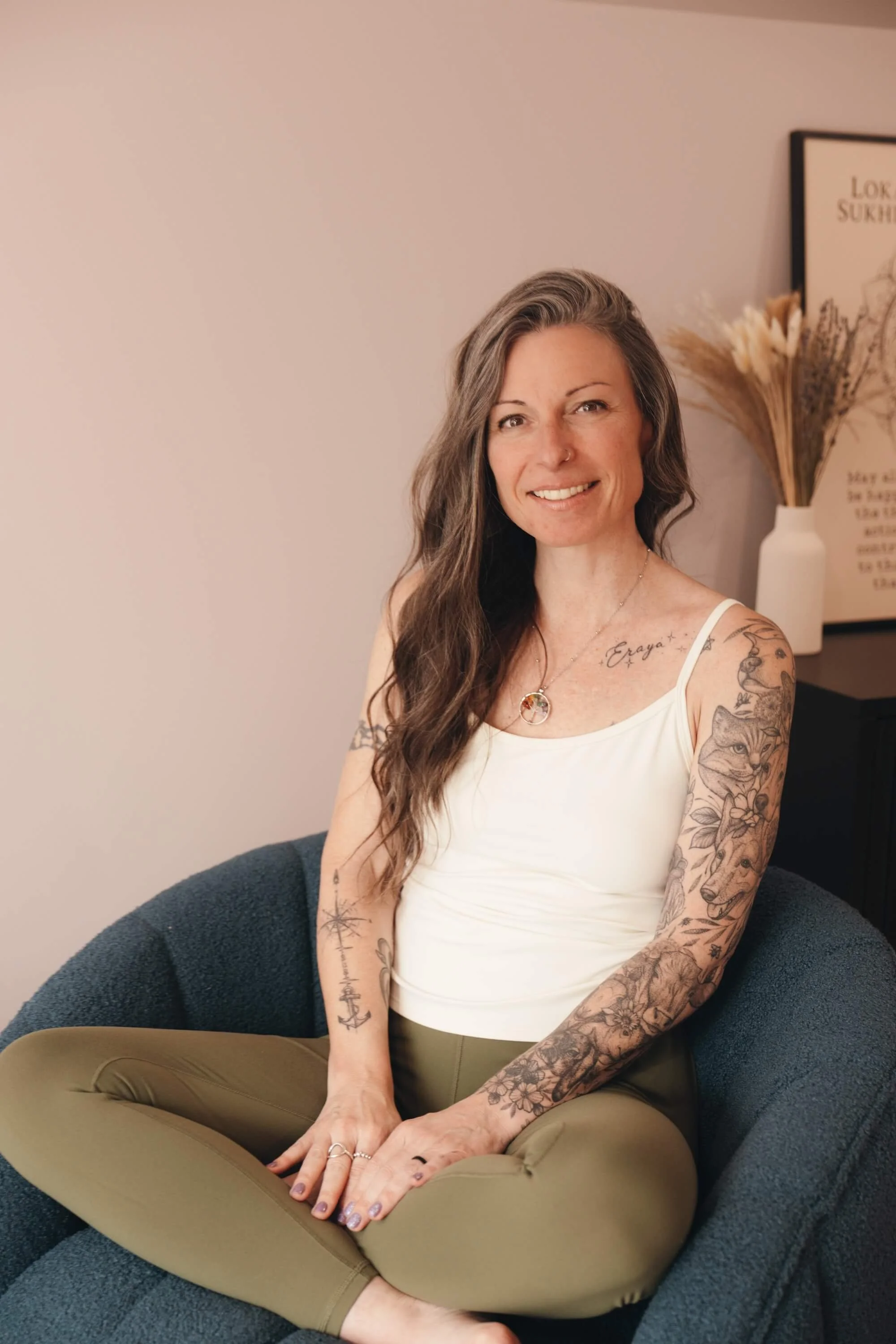A woman with long wavy hair and tattoos on her arms and chest, sitting cross-legged on a dark upholstered chair, smiling at the camera, in a room with a beige wall, a framed poster, and a white vase with dried flowers.
