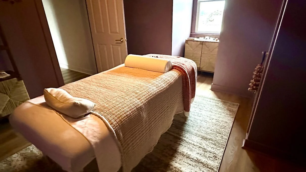 A massage room with a massage table covered in a beige blanket, with pillows and a bolster, next to a window, carpeted floor, and wooden furniture.