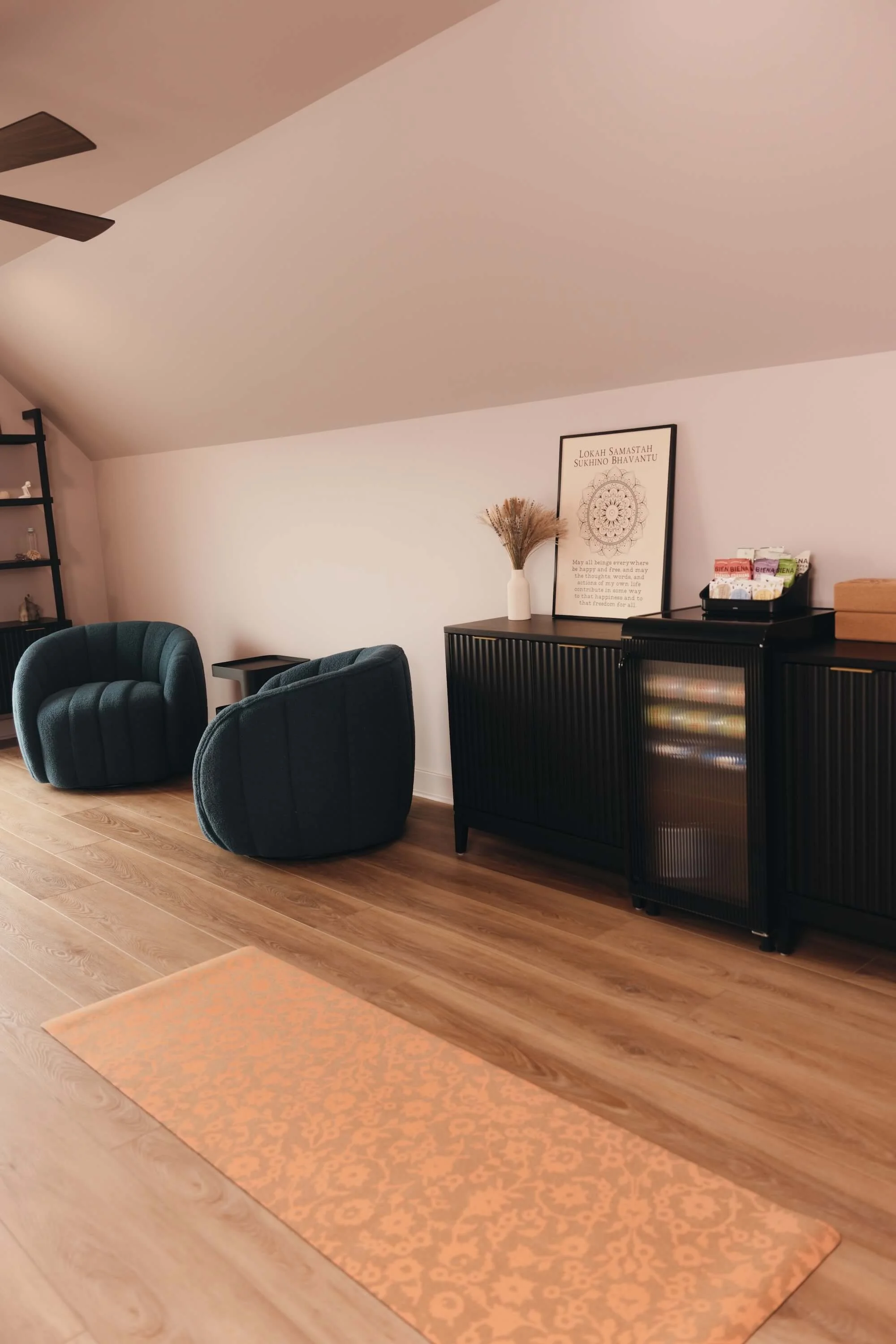 Studio with wooden flooring, black furniture, two dark teal armchairs, a black cabinet with decor and a framed quote, a pinkish rug, and a sliding door.