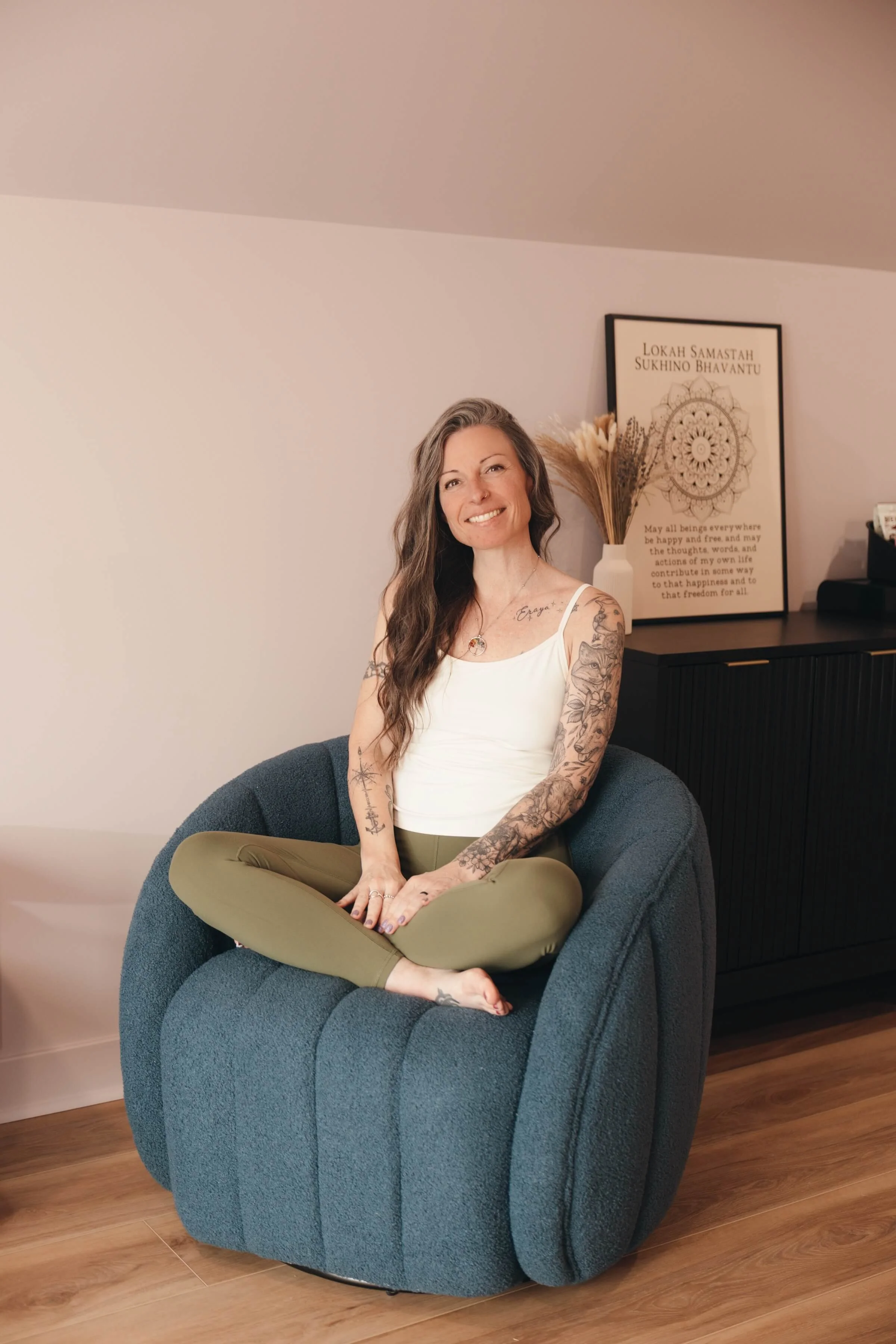 A woman sitting cross-legged on a blue armchair in a living room. She has long, wavy brown hair, tattoos on her arms, and is smiling at the camera. Behind her, a framed quote and a vase with dried flowers are visible on a black cabinet.