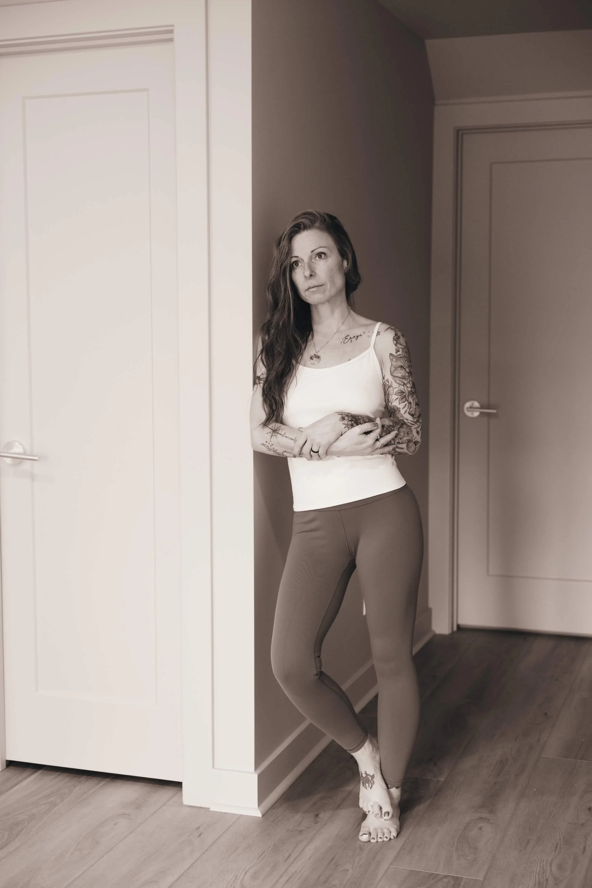 A woman with tattoos on her arms and shoulder, wearing a white tank top and beige leggings, leaning against a corner wall in a room with closed doors and wooden flooring.