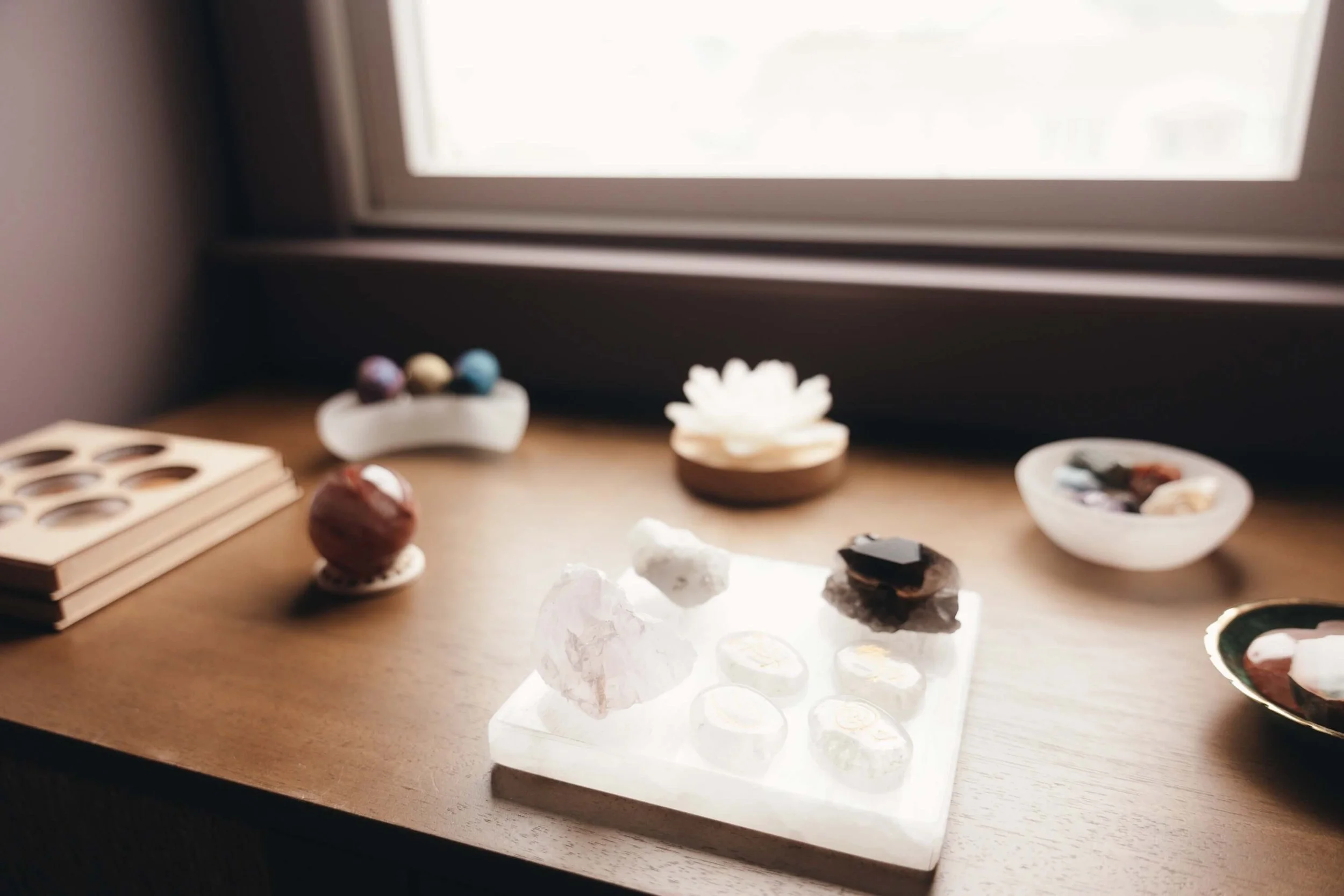 Close-up of a window sill with various stones and minerals displayed on a white stand and bowls, and a wooden puzzle or game with circular carved sections on a desk or table.