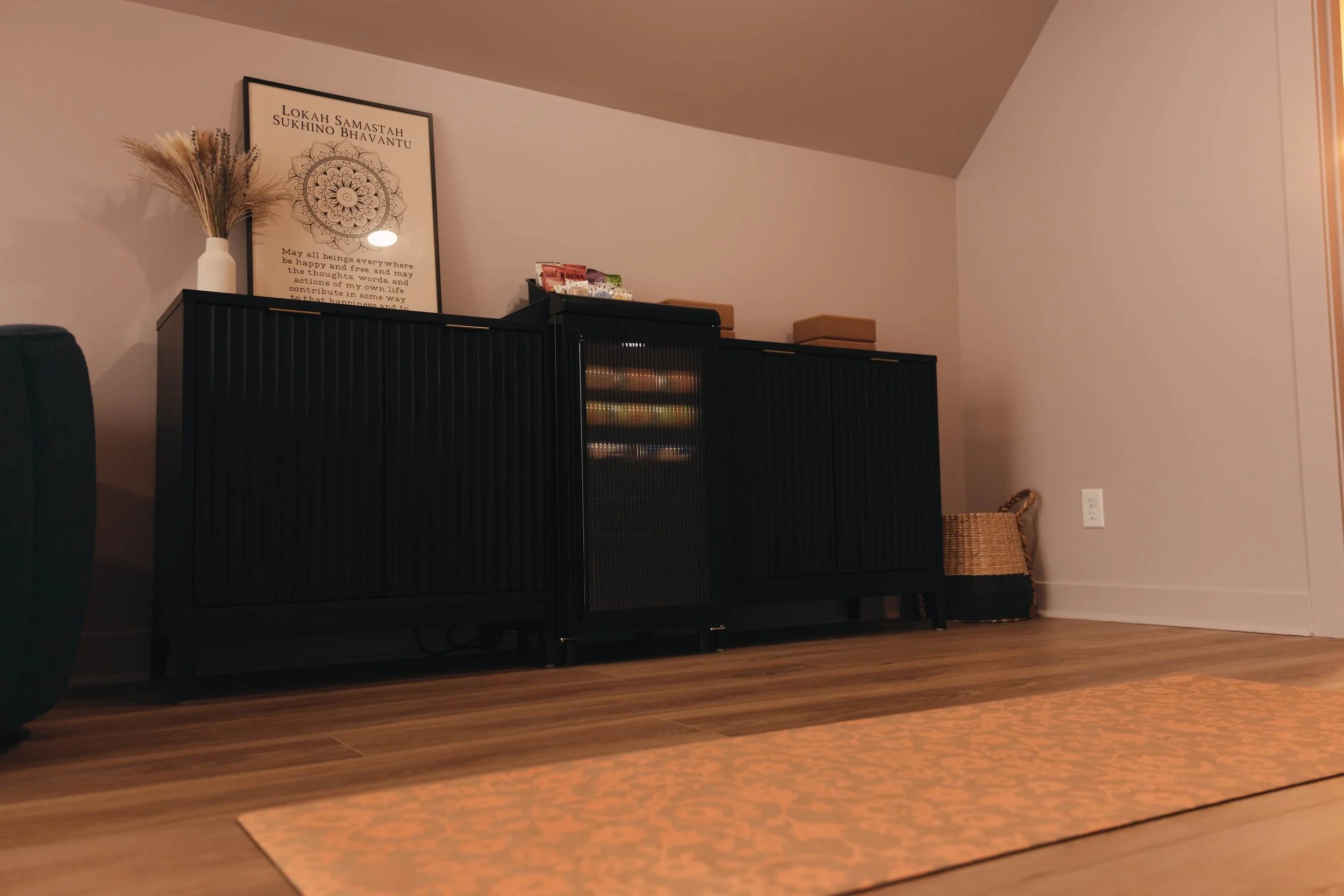A black sideboard with a framed decorative print, a white vase with wheat, snack packages, and a small box on top, against a beige wall with an angled ceiling.