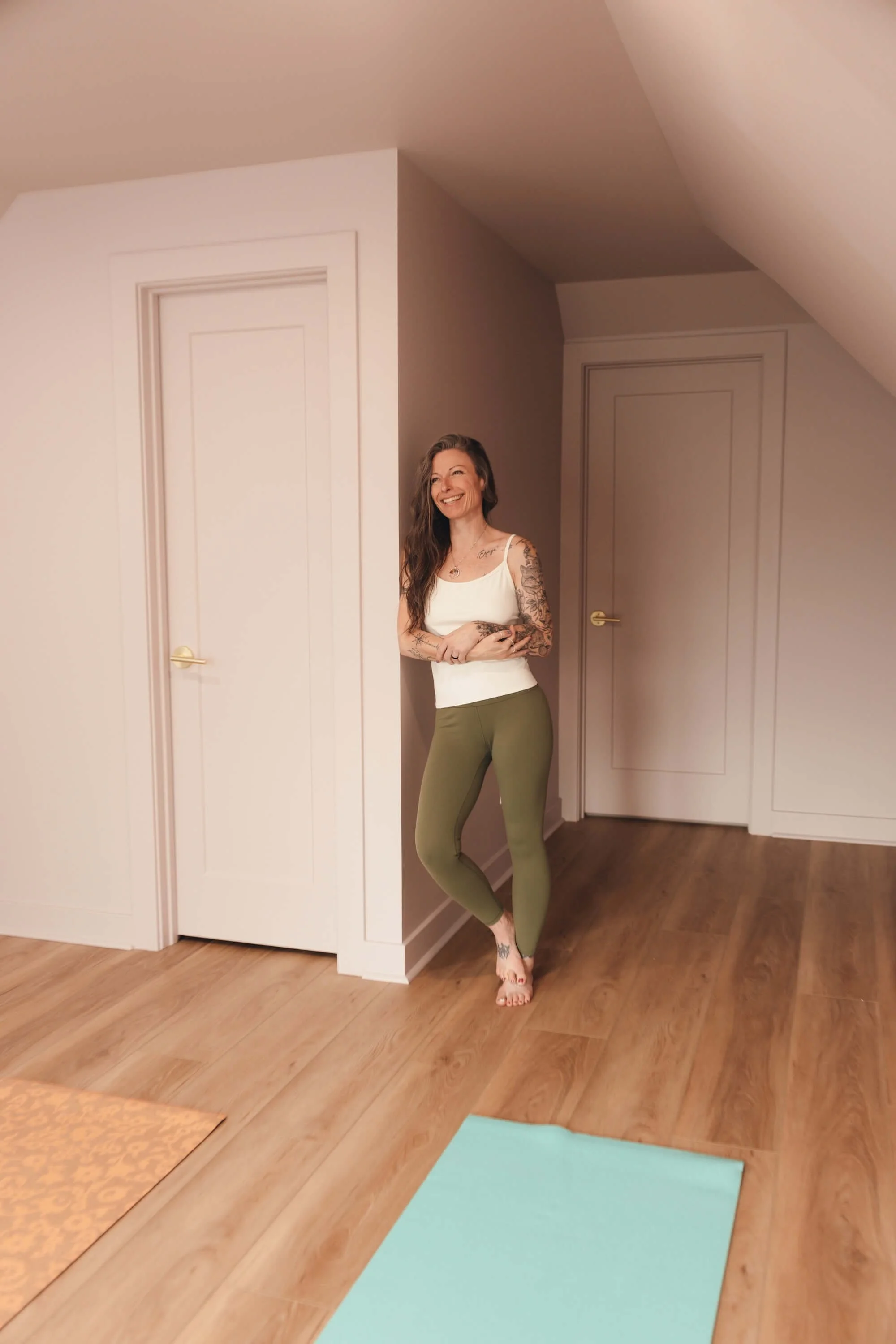 A woman with tattoos standing indoors near a corner wall, smiling, wearing a white tank top and green leggings, with yoga mats on the wooden floor.