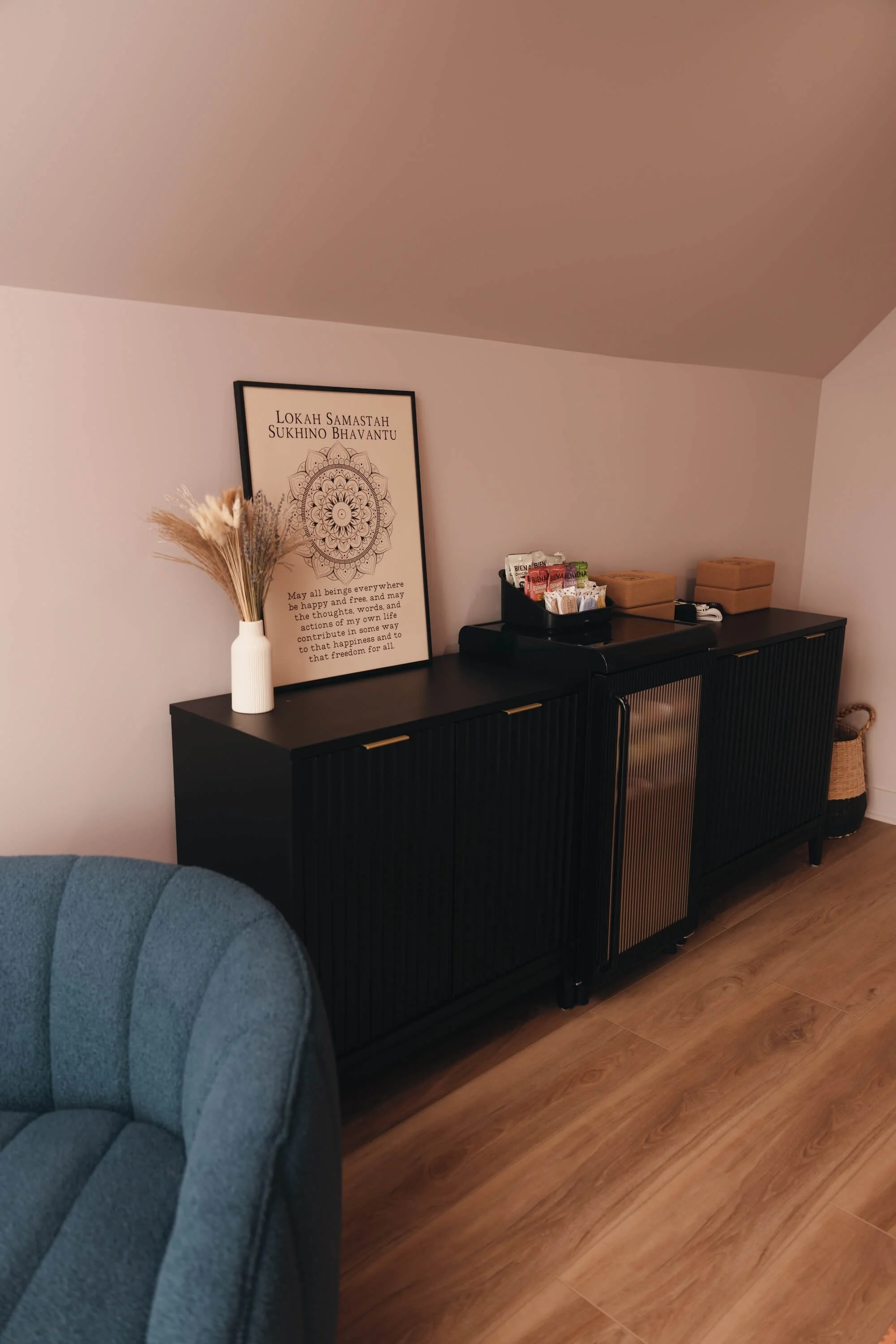 A black sideboard with a framed quote poster on the wall, a white vase with dried flowers, cups, and boxes on top, and a blue armchair partially visible in the foreground, all in a cozy room with wooden flooring.