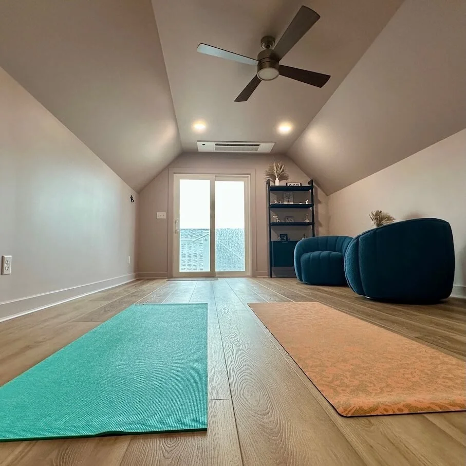A cozy attic living room with a ceiling fan, sliding glass door leading to a balcony, two blue armchairs, a black shelving unit, and two yoga mats on wooden flooring.