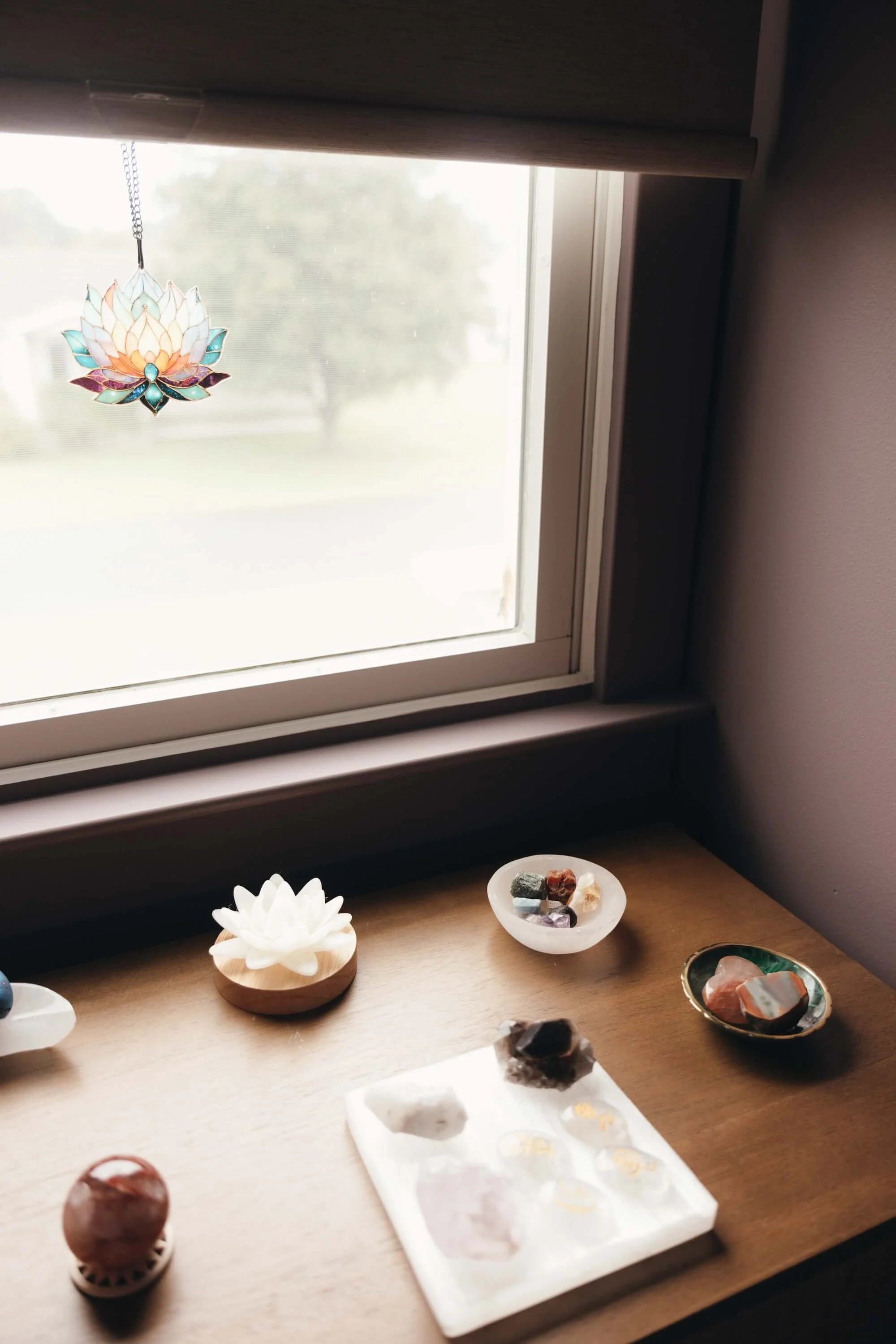 Second-floor window with a stained glass lotus leaf hanging decoration, a table with various mineral and gemstone specimens in bowls, and a white lotus flower candle holder. The scene is lit by natural light from outside, with a view of trees and cloudy sky.