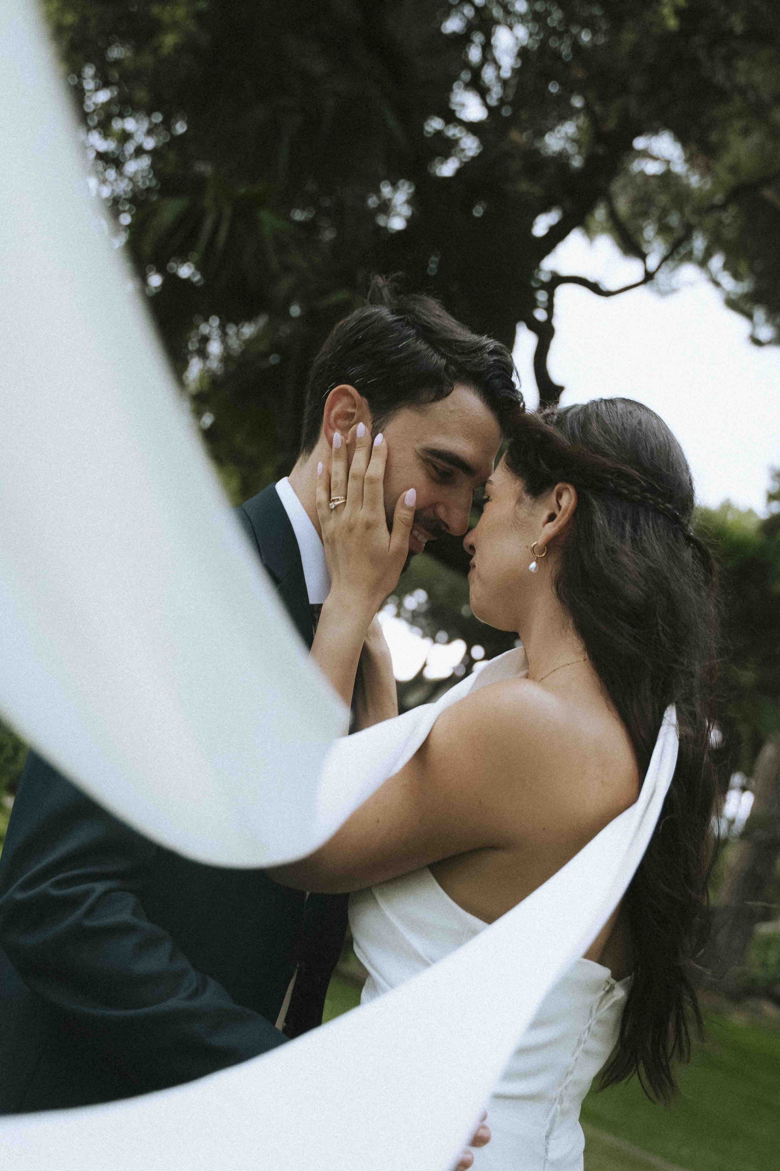 A bride and groom in an intimate moment outdoors, with their foreheads and noses touching, and eyes closed. They are embracing each other, surrounded by green trees and an overcast sky.