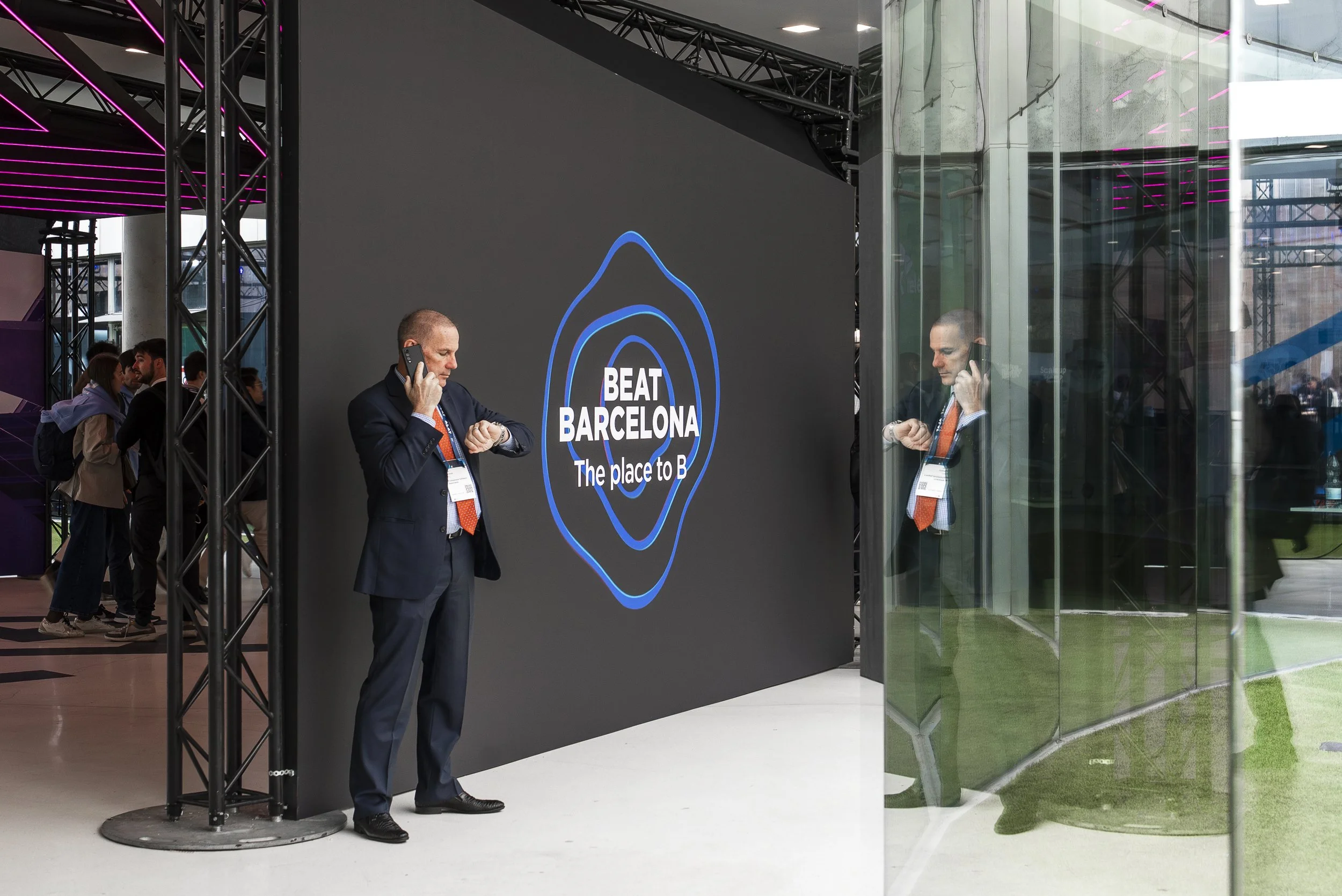 A man in a suit and orange tie standing indoors, talking on a cell phone, with a large digital screen behind him displaying the text 'BEAT BARCELONA The place to B' with a blue outline of a location pin. Several people are in the background, and the area has a modern, industrial design with metal beams and glass walls.