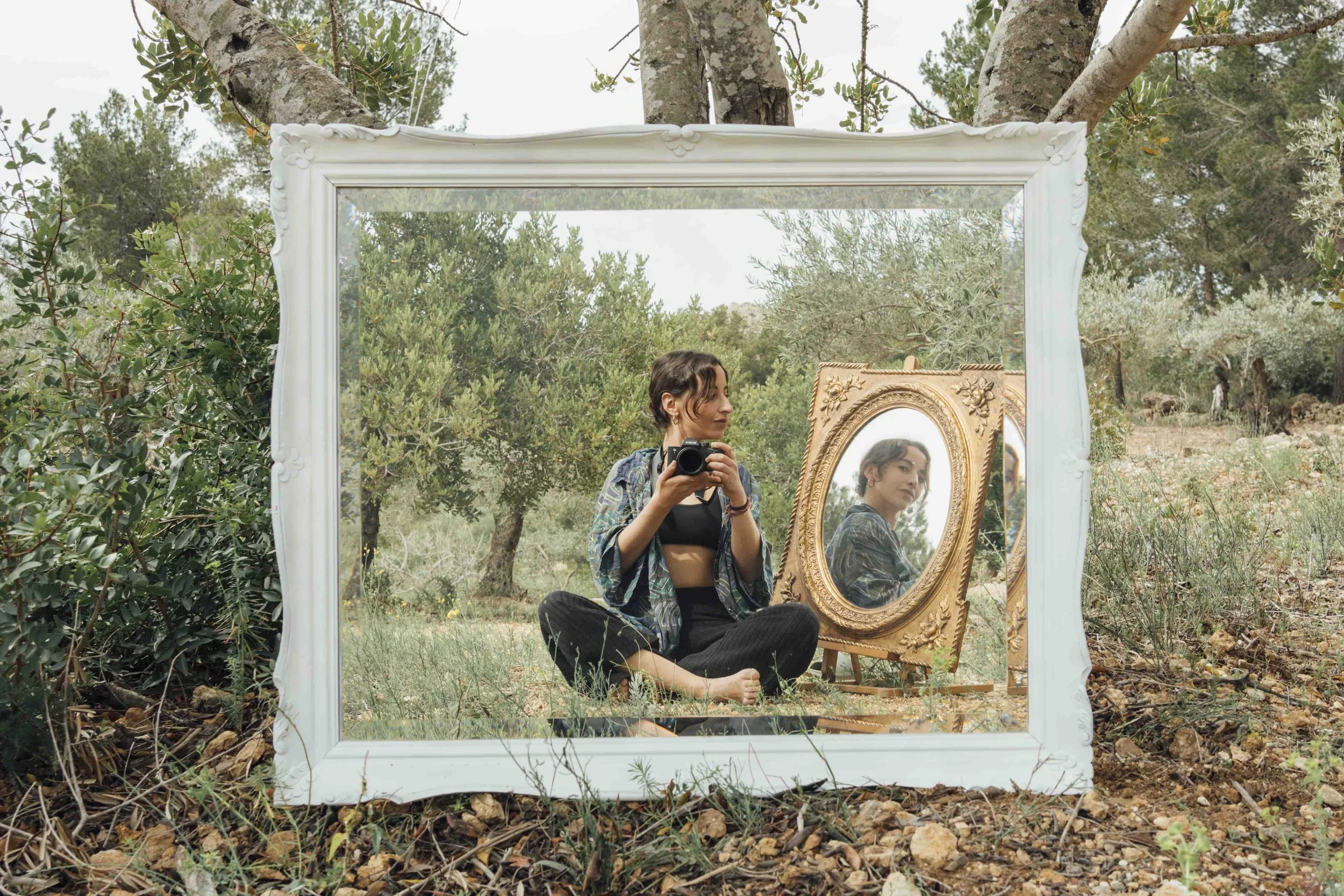 A woman sitting outdoors in front of a white mirror frame and a gold ornate mirror, holding a camera, with trees and bushes surrounding her.