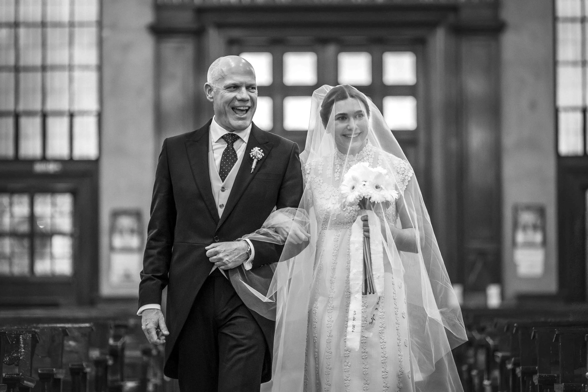 A black and white photo of a wedding ceremony inside a church. The groom, a bald man in a black suit and polka dot tie, is smiling as he walks down the aisle. The bride, wearing a lace wedding dress and veil, holds a bouquet and smiles as well.