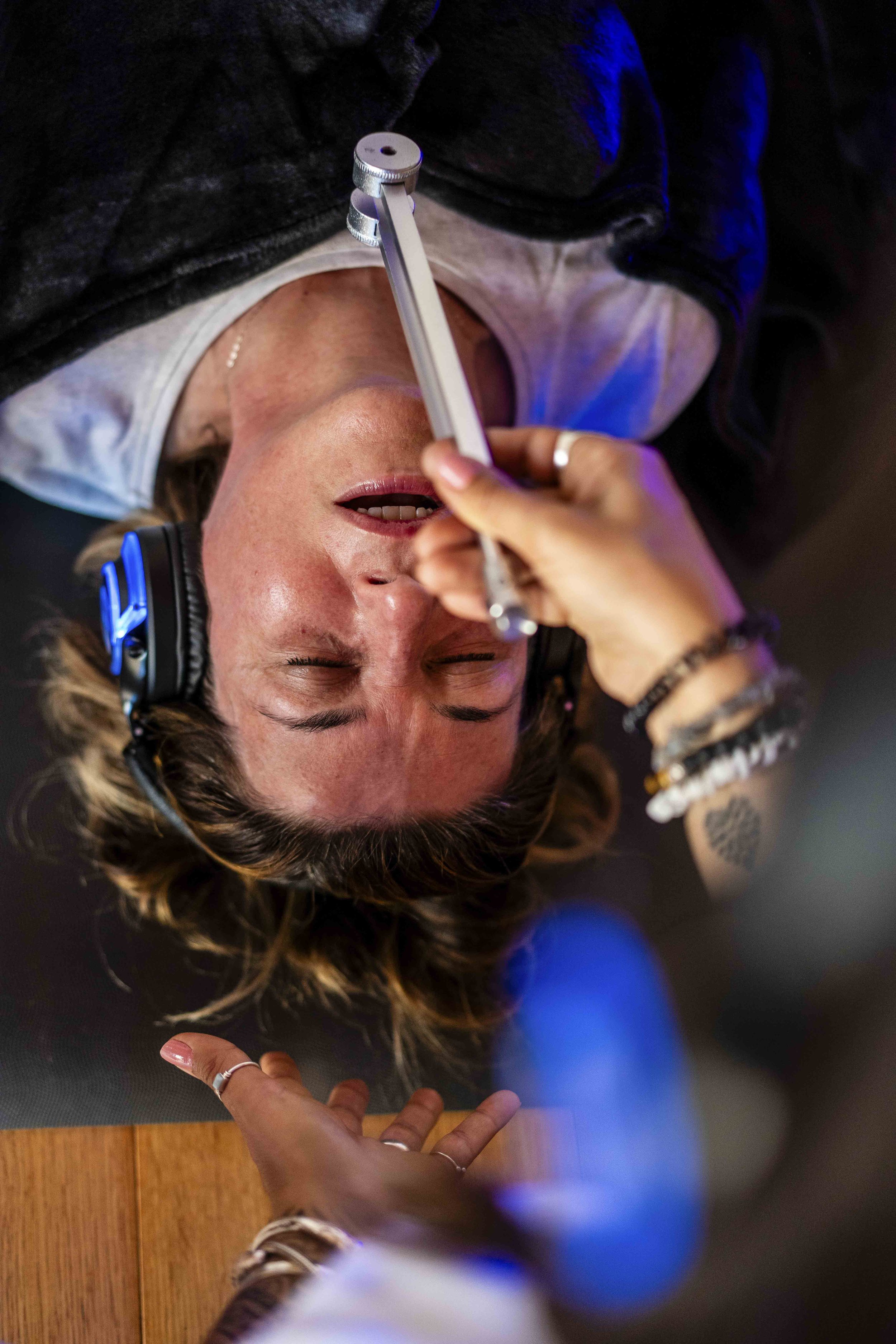 Person lying on their back with closed eyes, wearing headphones, with a feathered bracelet and rings on their fingers, holding a pencil or stylus in one hand and resting on a wooden surface.