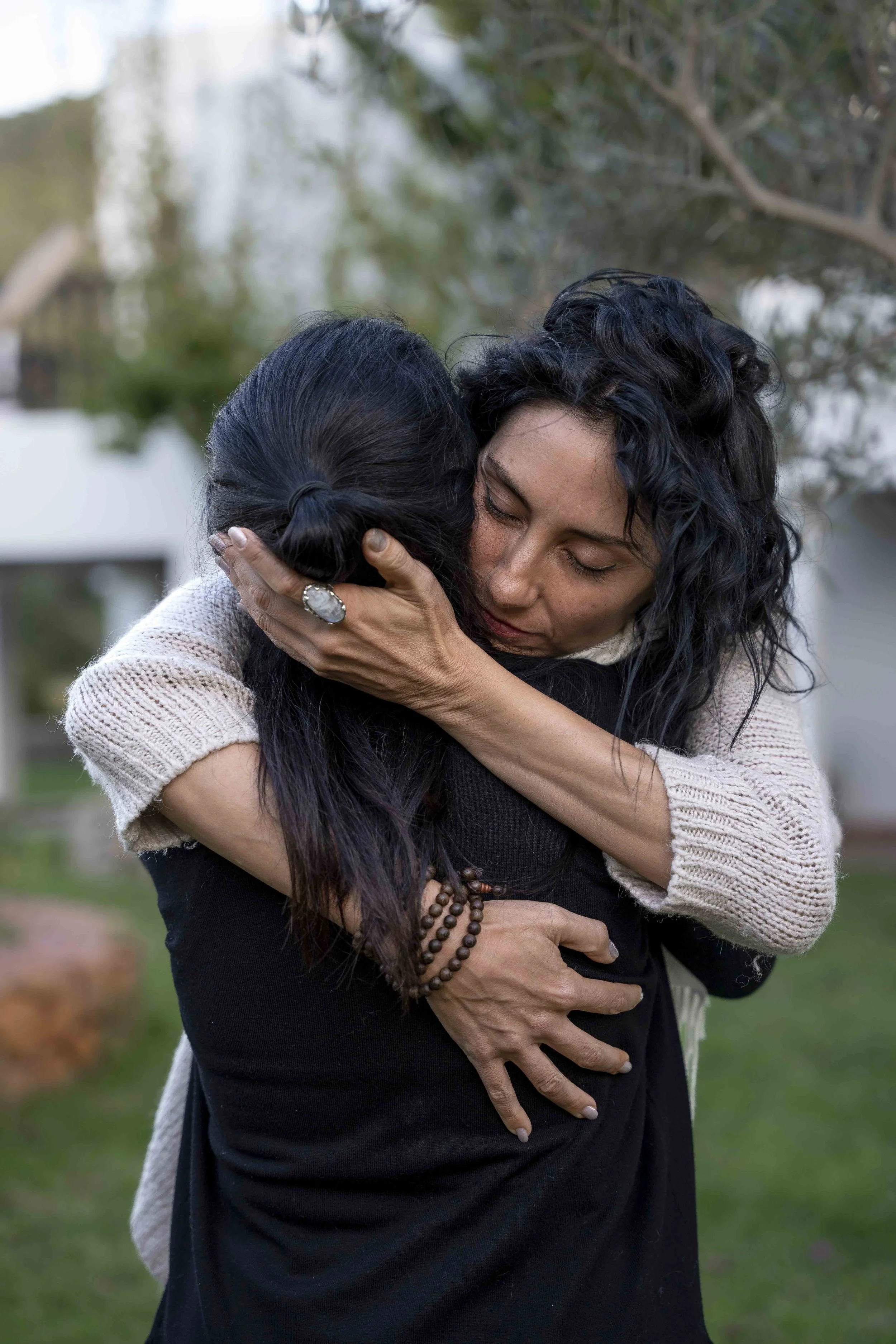 Two women hugging outdoors, one with curly black hair and the other with straight black hair tied in a bun, surrounded by trees and greenery, conveying emotion and connection.