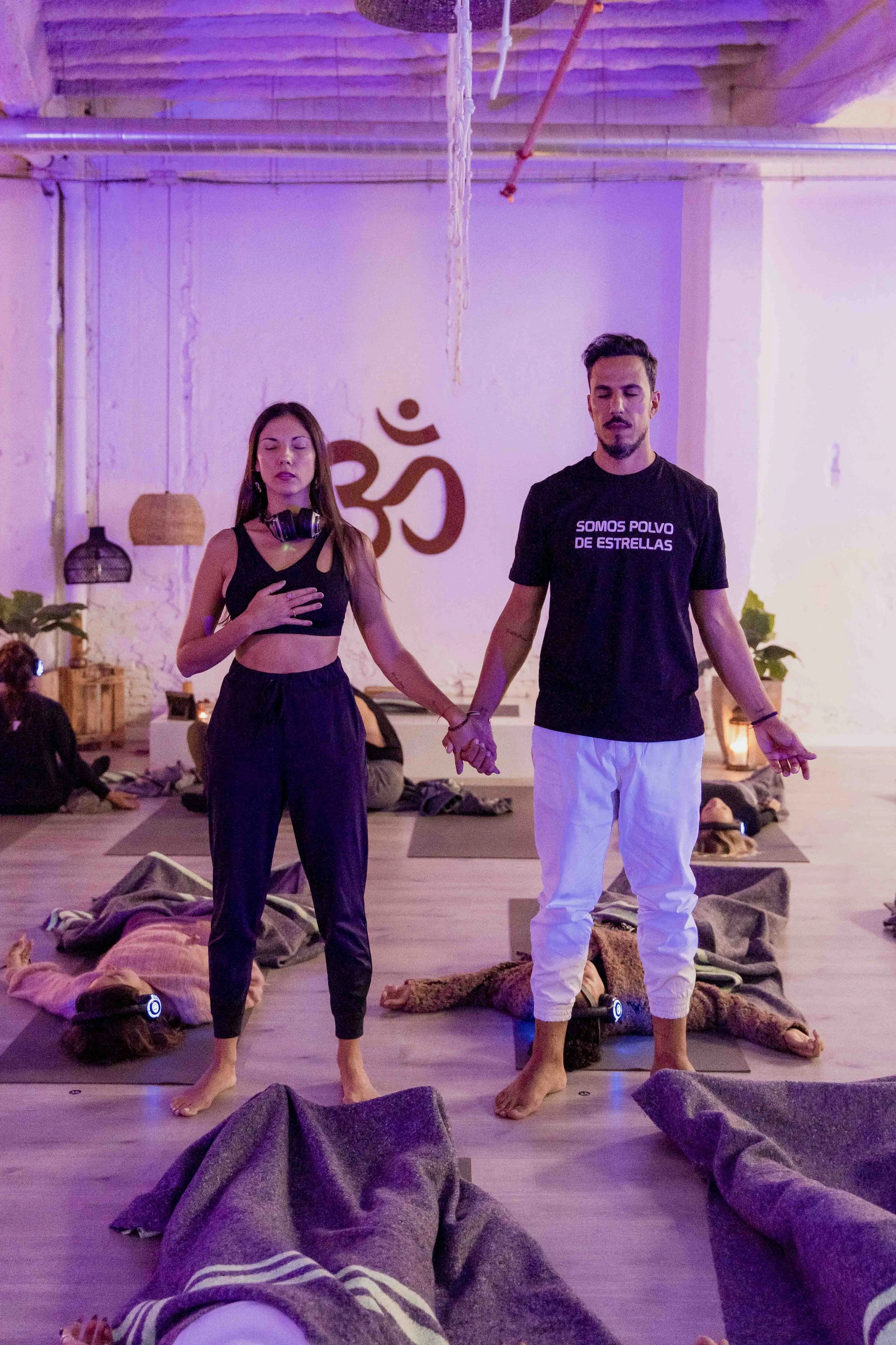 A group of people practicing yoga or meditation in a dimly lit studio with purple and pink lighting, some lying on mats and some standing with hands in prayer positions.