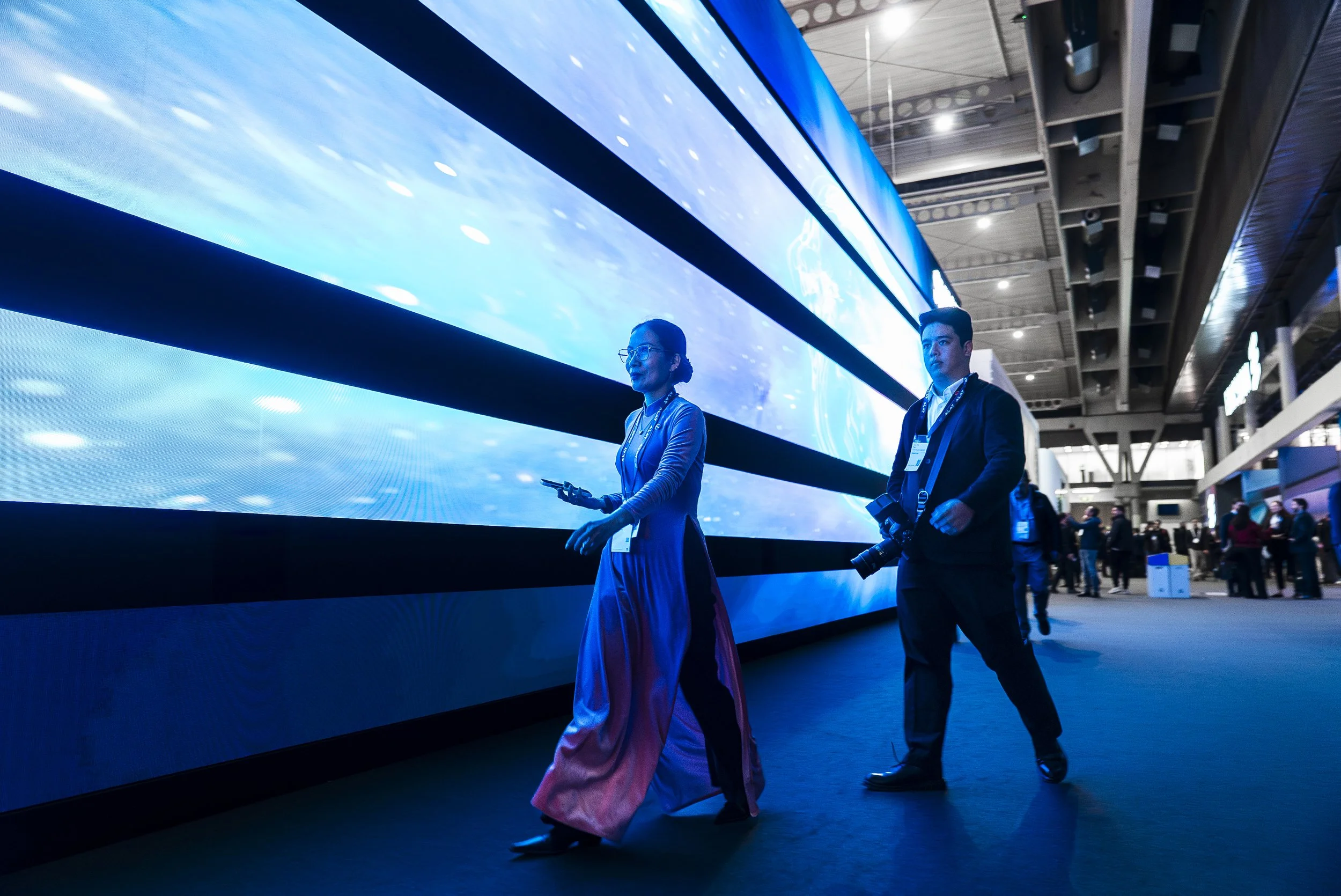 People walking past a large digital display at an indoor event or conference, illuminated with bright blue colors and cyber images.