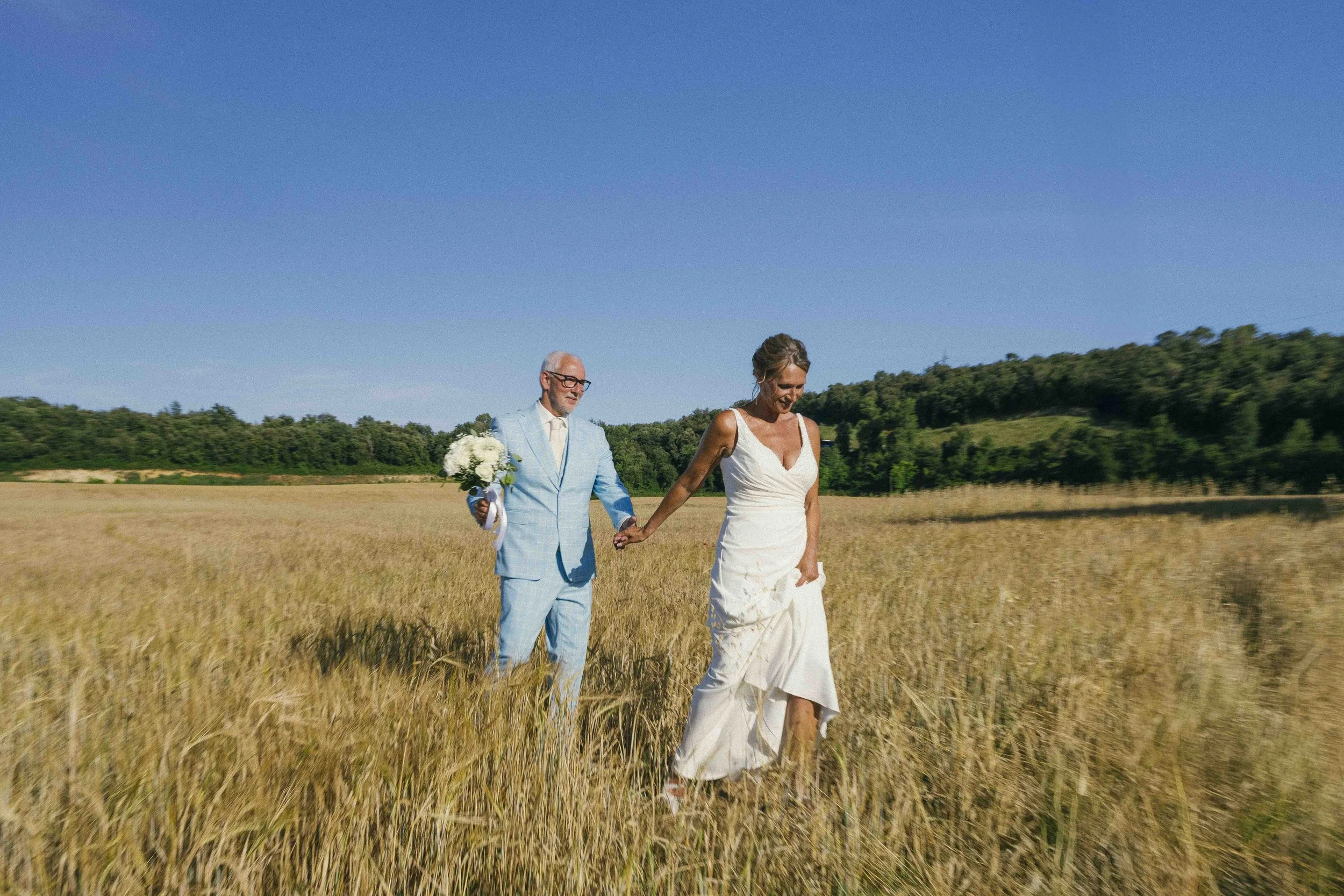 A bride and groom walking hand in hand through a field of tall grass under a clear blue sky on their wedding day.