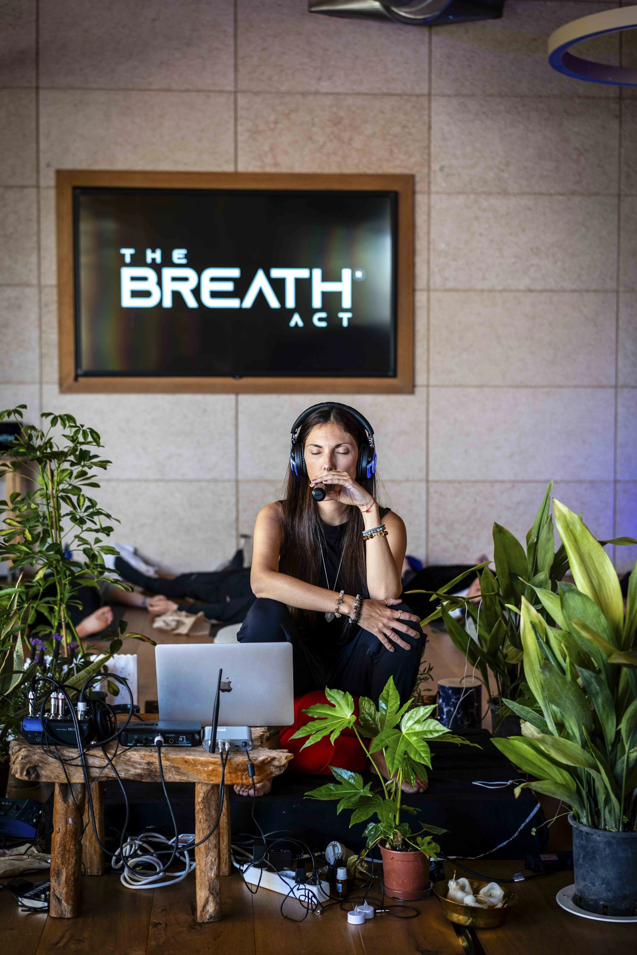 A woman with long brown hair, wearing headphones, sitting on the floor with her eyes closed and hand over her mouth, surrounded by plants, electronic equipment, and a laptop, with a TV screen in the background displaying 'The Breath Act'.