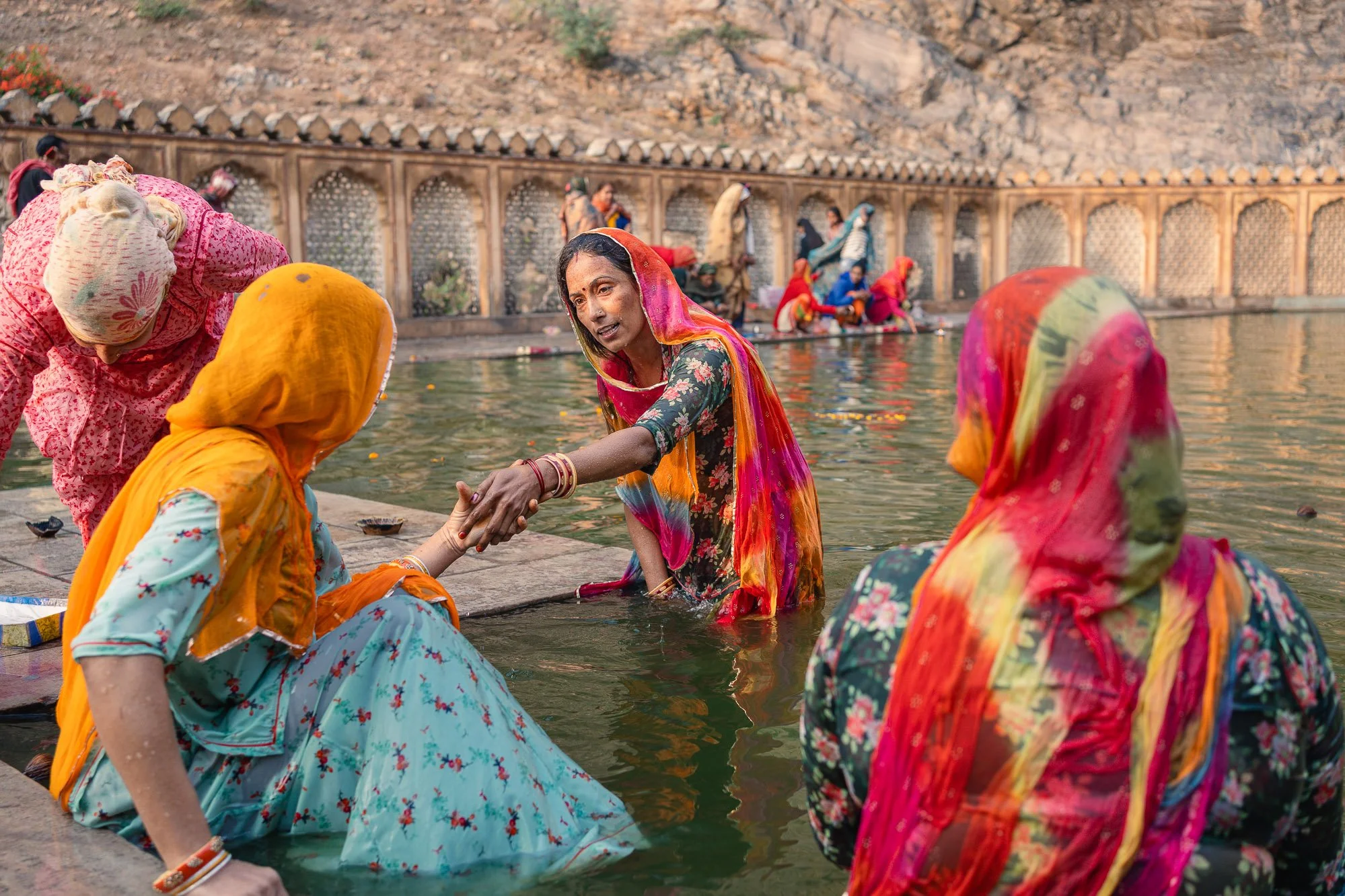 Women in colorful traditional Indian clothing performing a religious ritual in a river, with onlookers in the background and a stone wall along the riverbank.
