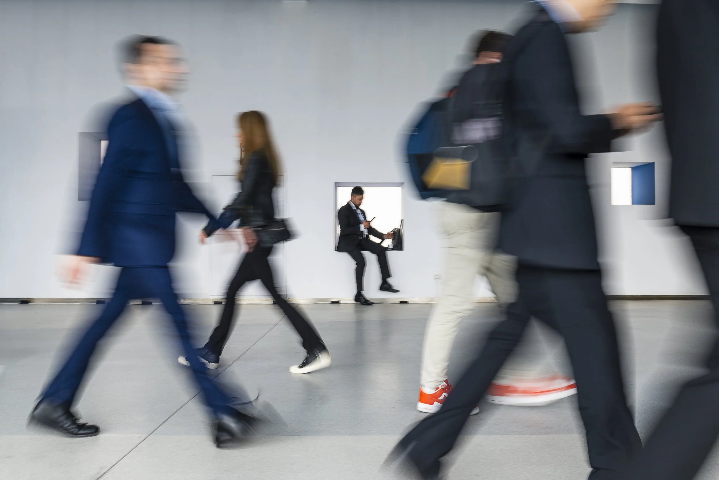 Blurred motion of people walking in an indoor space, with one person sitting and using a laptop against a white wall with small square windows.