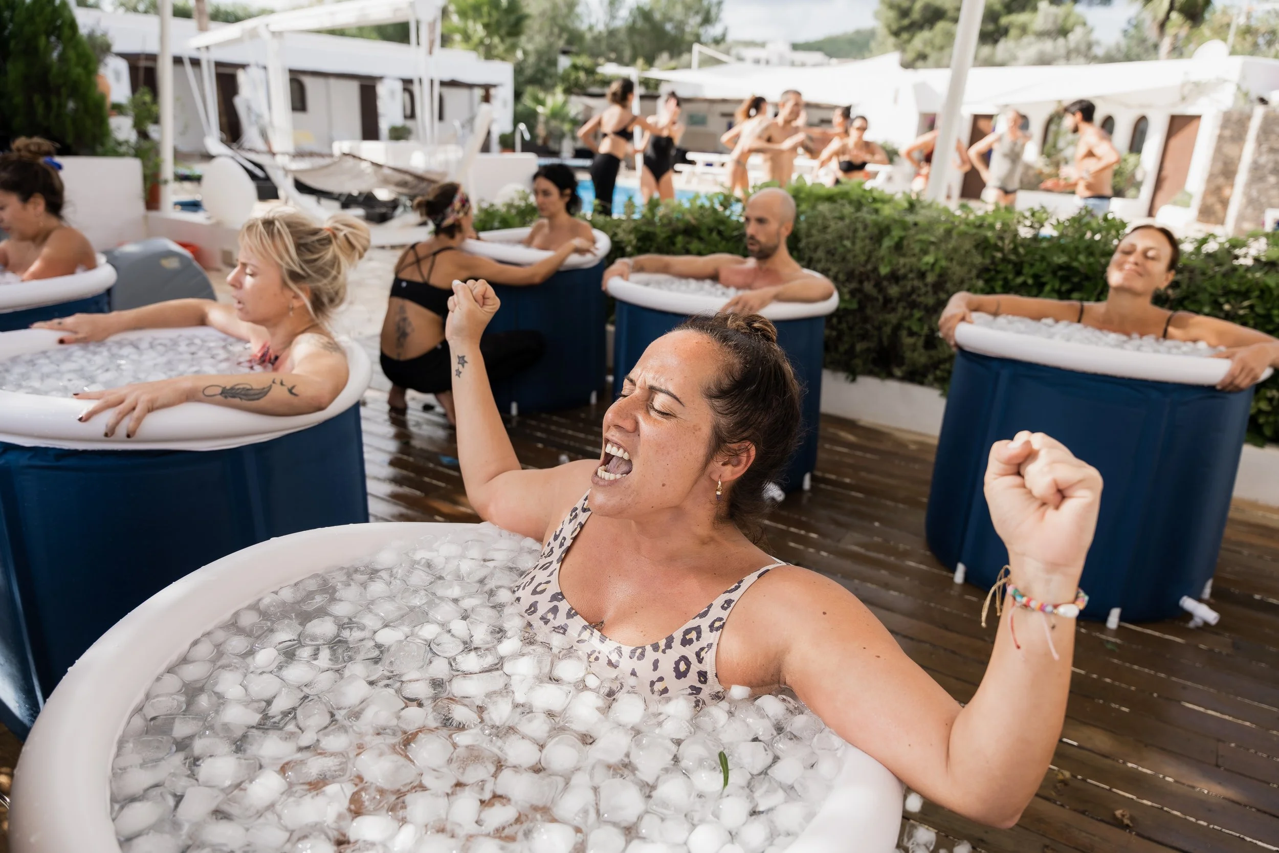 People relaxing in small inflatable pools filled with ice on a wooden deck, with a group of women in the background at a social gathering.