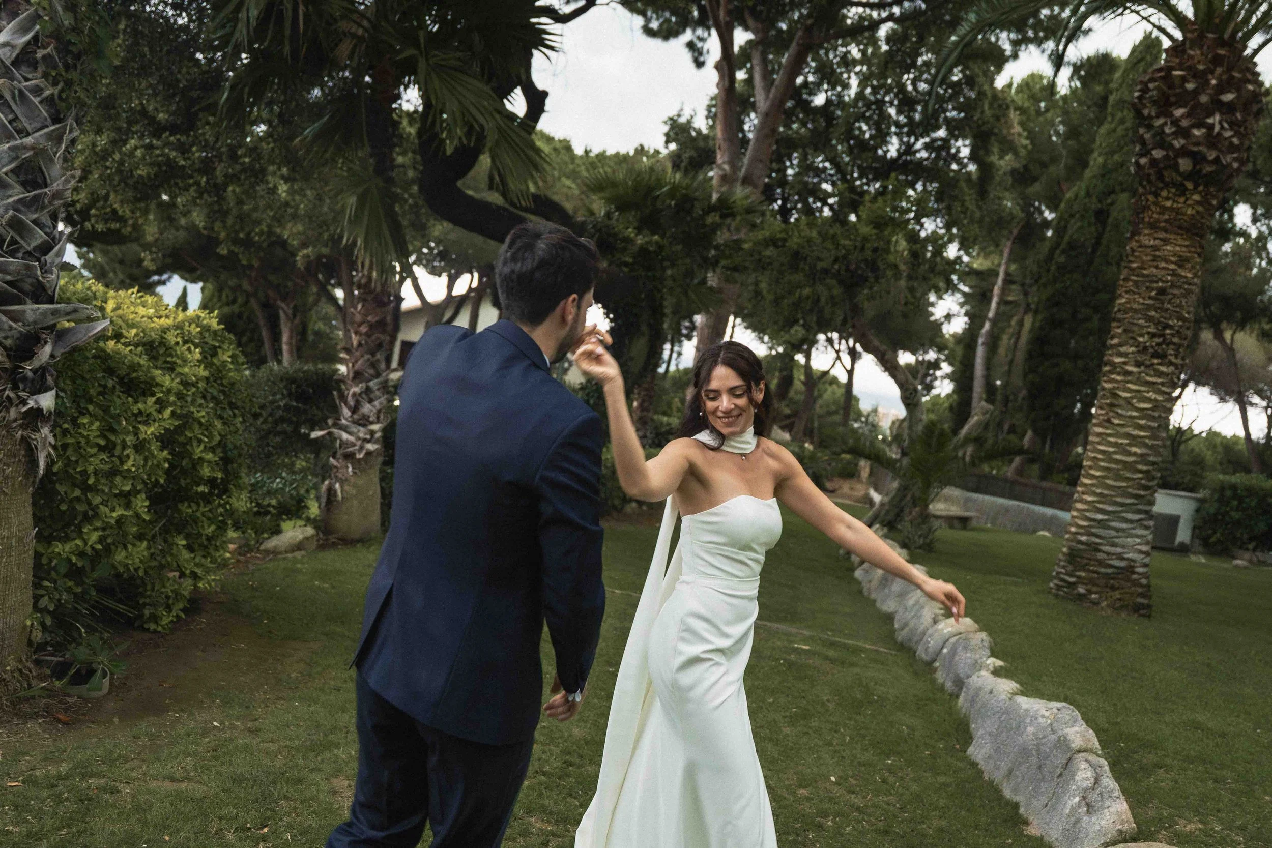 A man and a woman holding hands and dancing outdoors among trees and greenery, the woman in a white dress and the man in a dark suit.