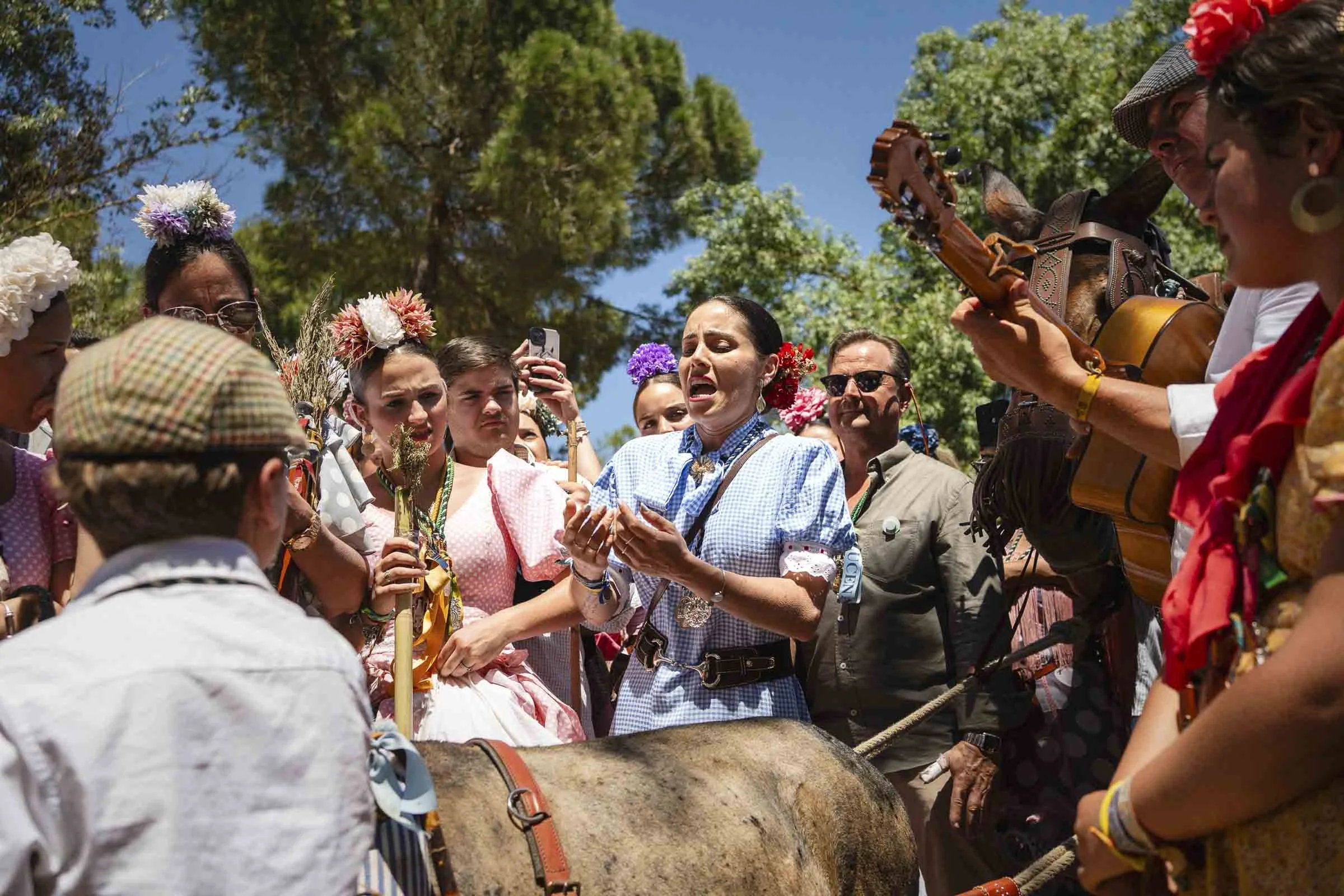 A group of people participating in a traditional Mexican celebration outdoors, dressed in colorful clothing and flower headbands, gathered around a horse, with a woman singing or speaking into a microphone and a woman playing the guitar.