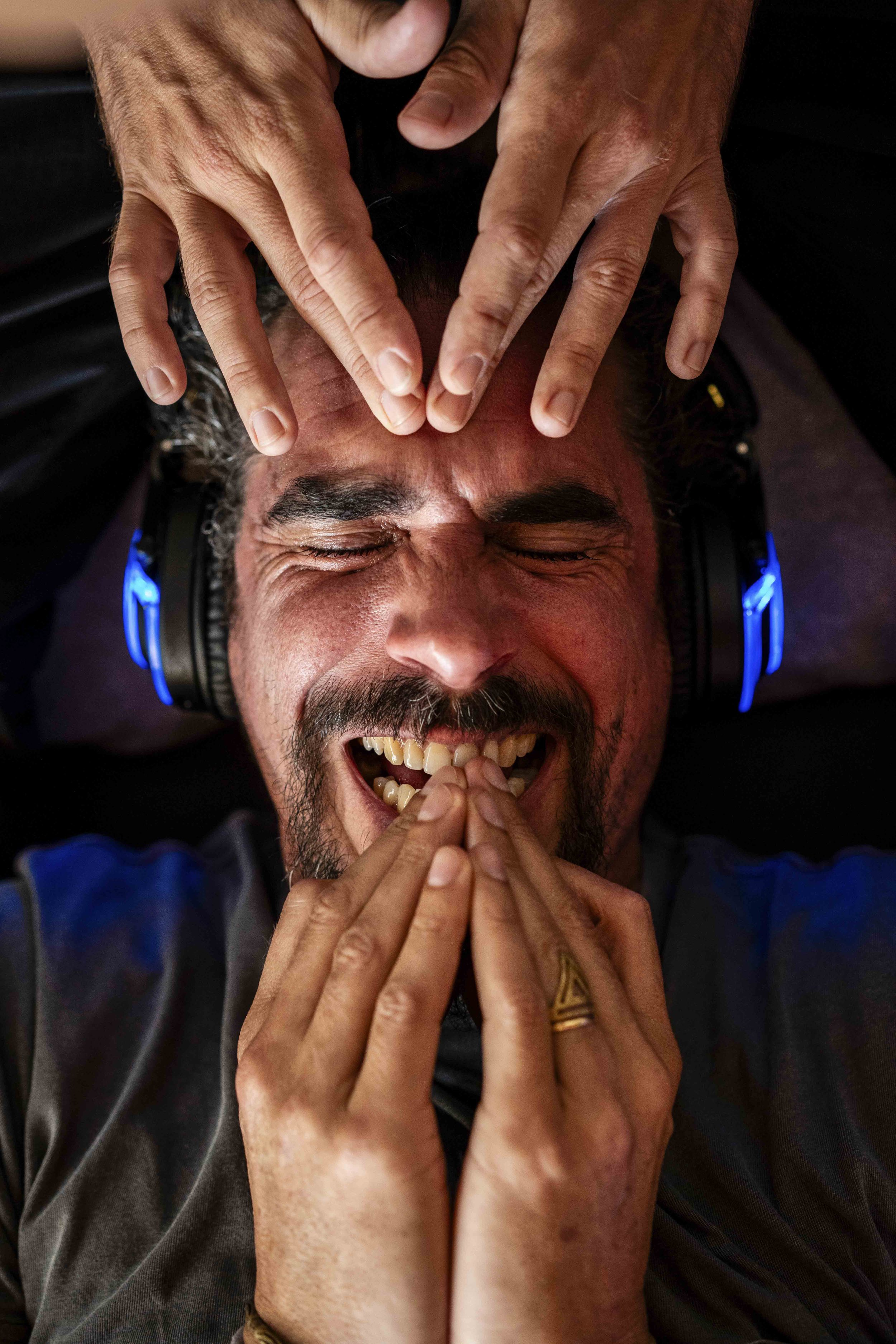 A person with a beard and closed eyes is smiling widely, wearing headphones, with hands placed on their forehead and chin, fingers interlocked, and a ring on their finger.