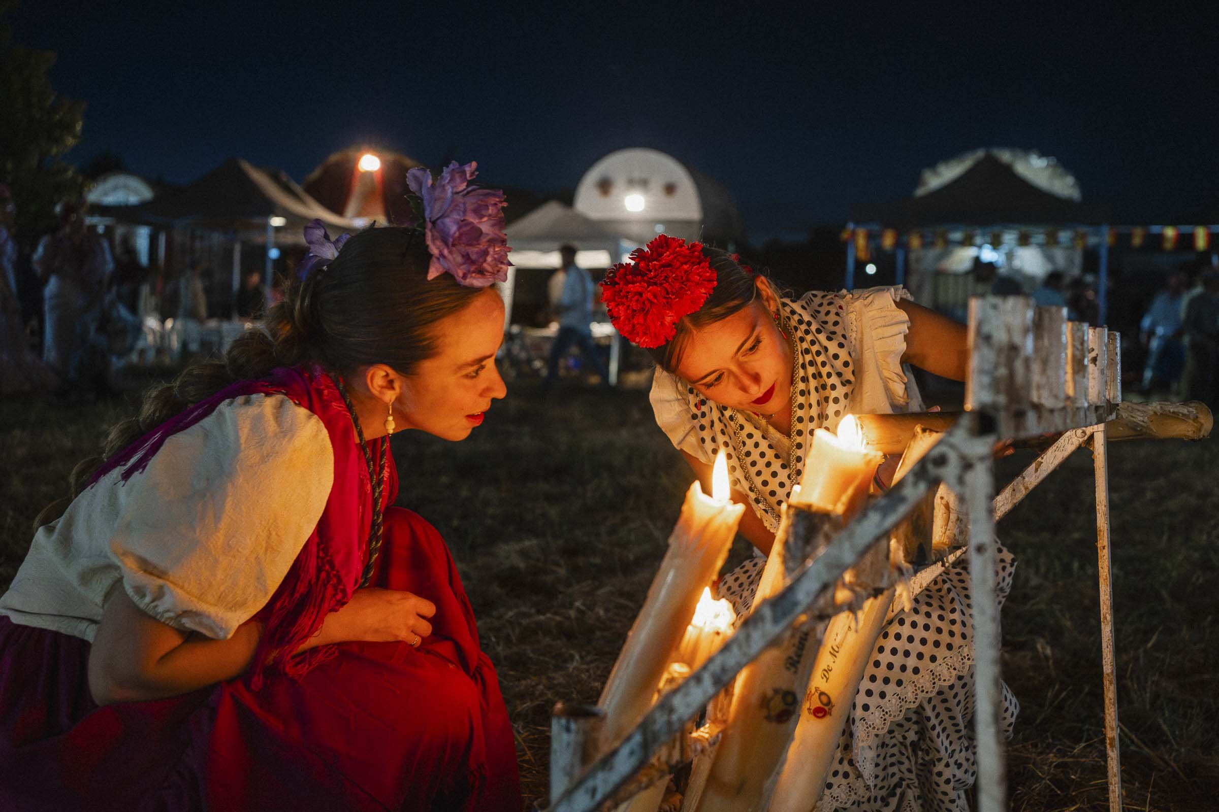 Two women dressed in traditional Mexican attire, with flowers in their hair, lighting candles at an outdoor event at night. In the background, tents and a white structure are visible under a dark sky.