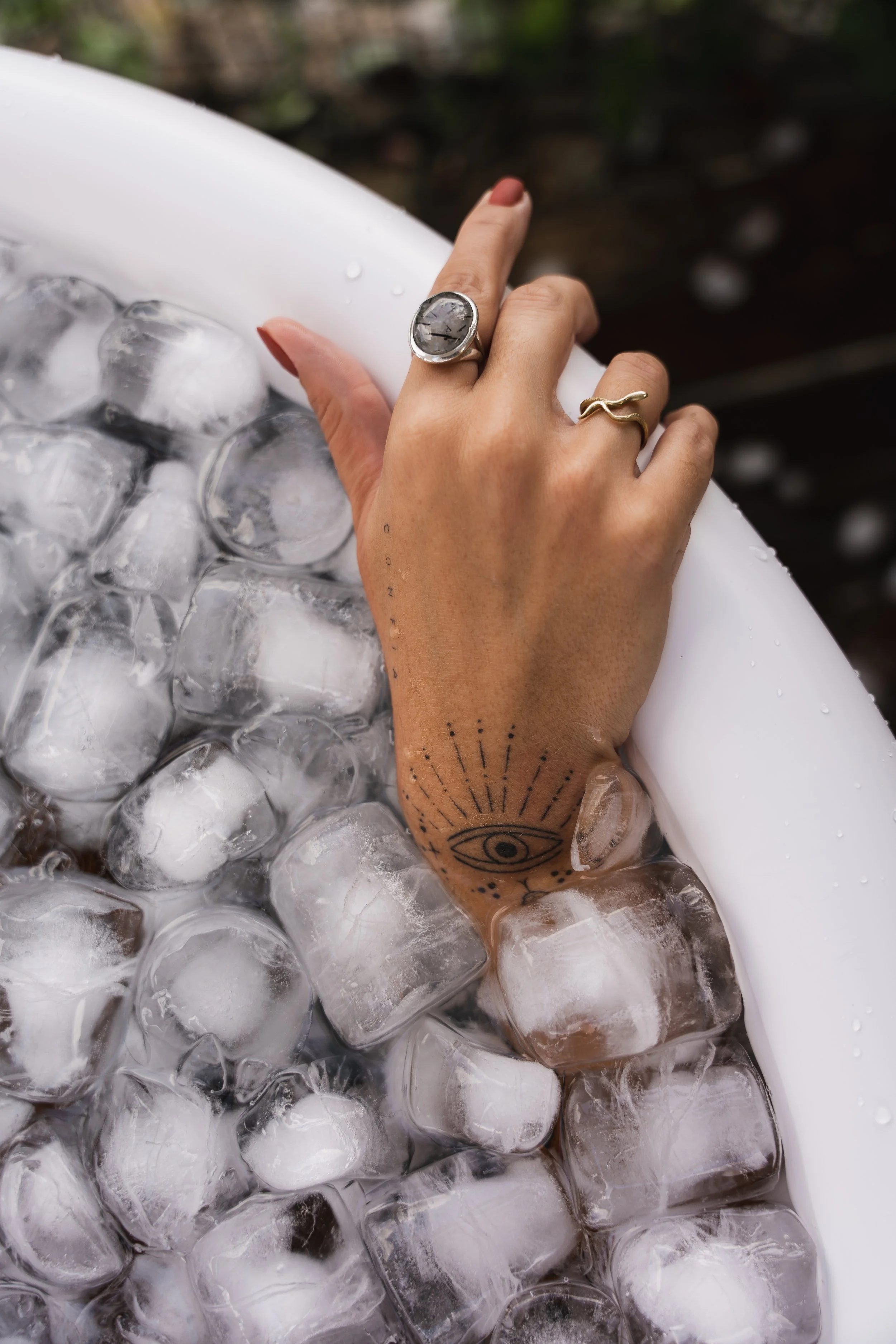 A person's hand with rings and tattoos resting on ice cubes inside a white container.