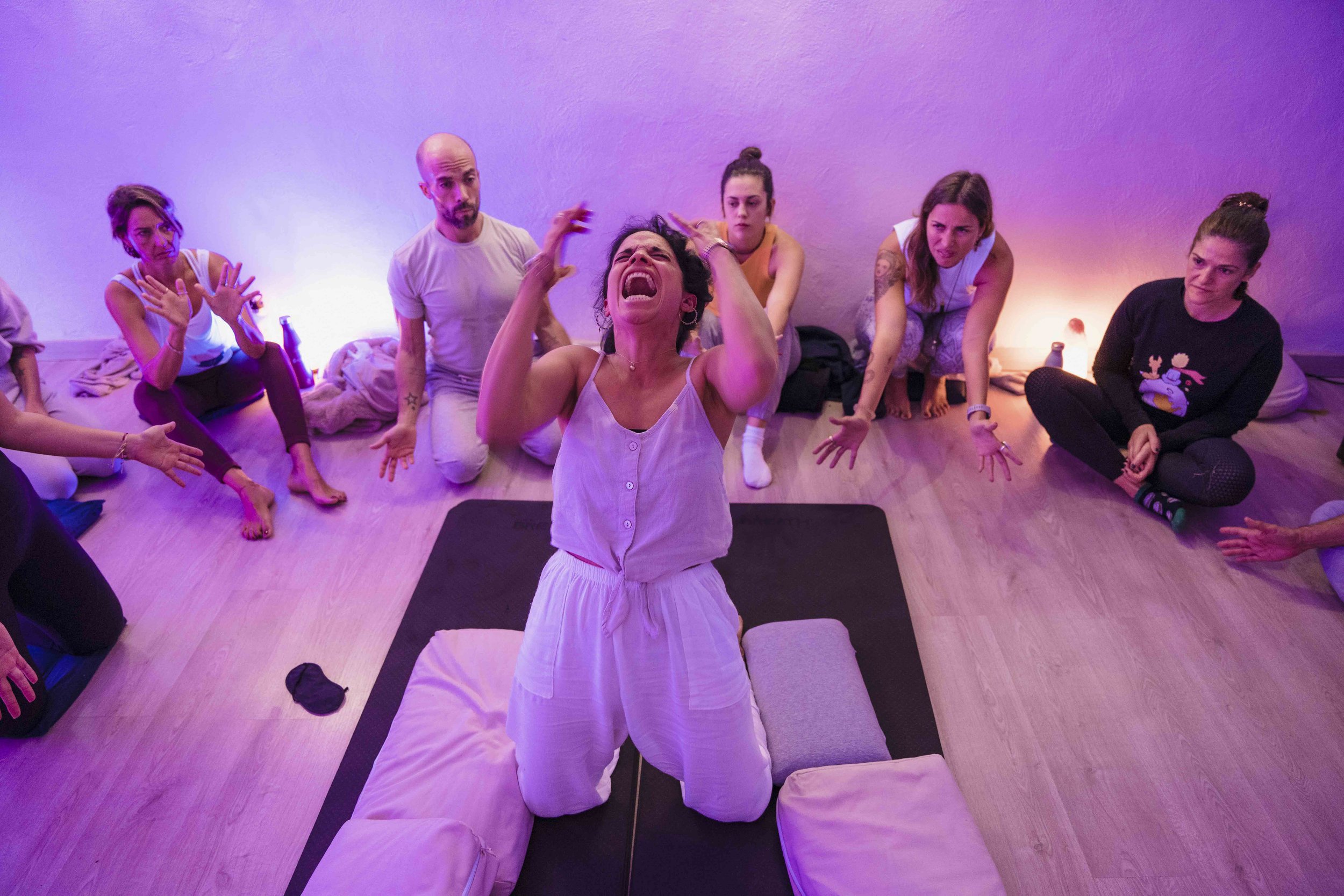 A group of people sitting on the floor in a room with purple and pink lighting, with one woman in a white dress kneeling in front, appearing to be speaking or yelling passionately.