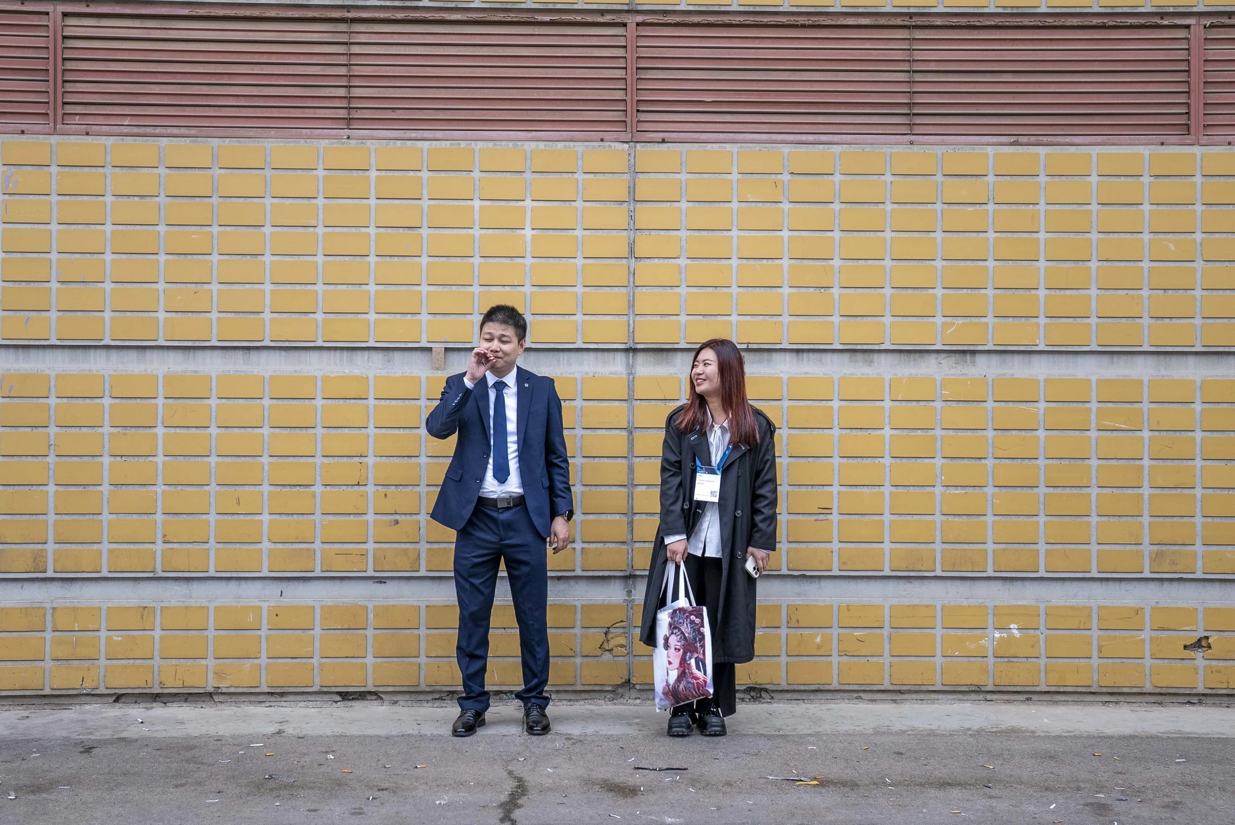 Two people, a man and a woman, stand in front of a yellow brick wall with red vent panels at the top. The man is wearing a navy suit and tie, has short black hair, and is smiling while touching his nose. The woman has long reddish-brown hair, is wearing a black jacket, a white shirt, chocolate sneakers, and is holding shopping bags, smiling while looking at the man.
