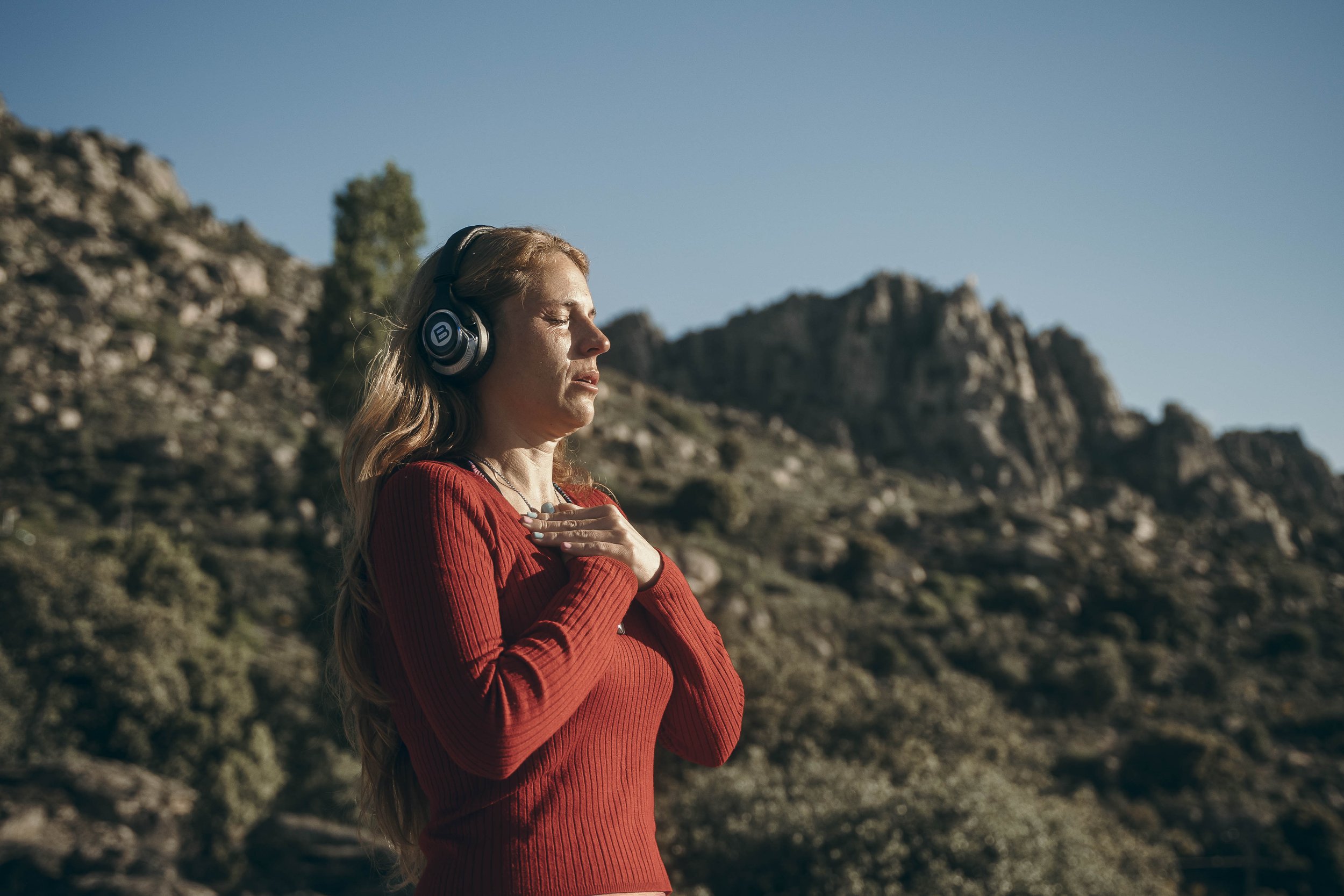 A woman with long blonde hair wearing a red sweater and headphones, standing outdoors with rocky hills and a clear blue sky in the background, her eyes closed and hands over her chest.