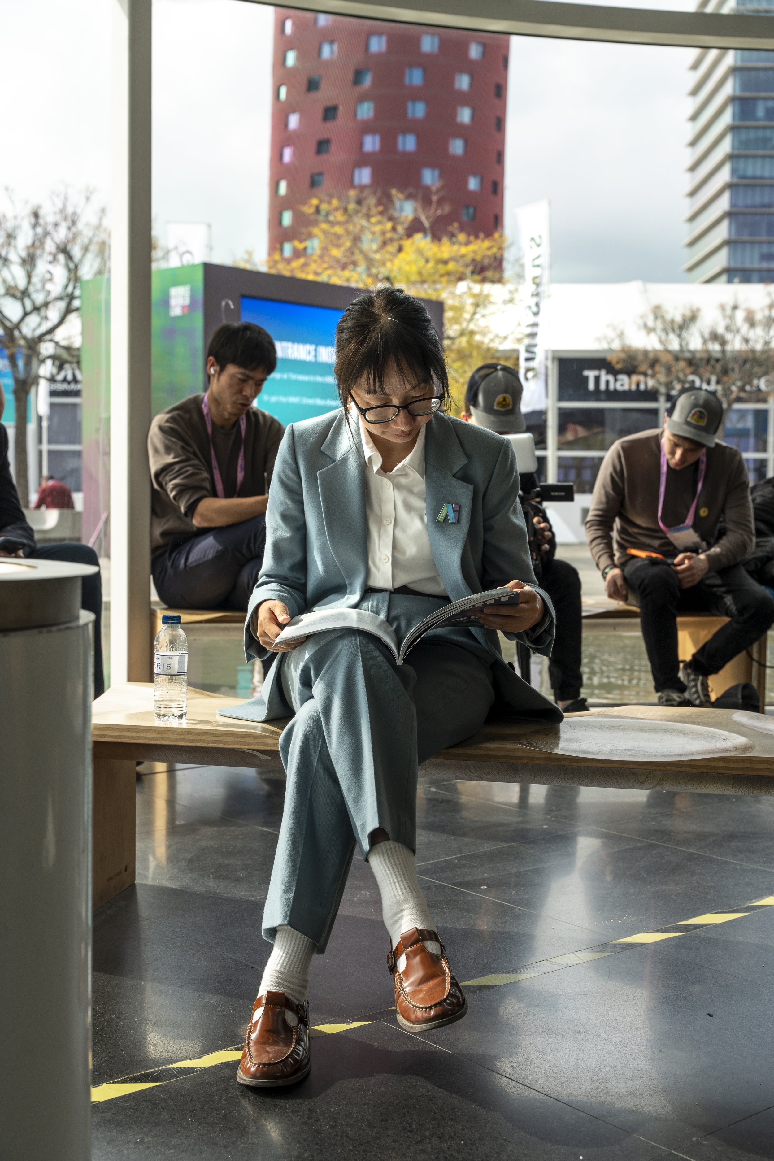 A woman in glasses, a gray suit, and white socks is sitting on a wooden bench, looking at a magazine. She has brown shoes. Two men sit behind her, one with a cap and a lanyard, and another with headphones, in an outdoor urban setting with modern buildings and digital screens.