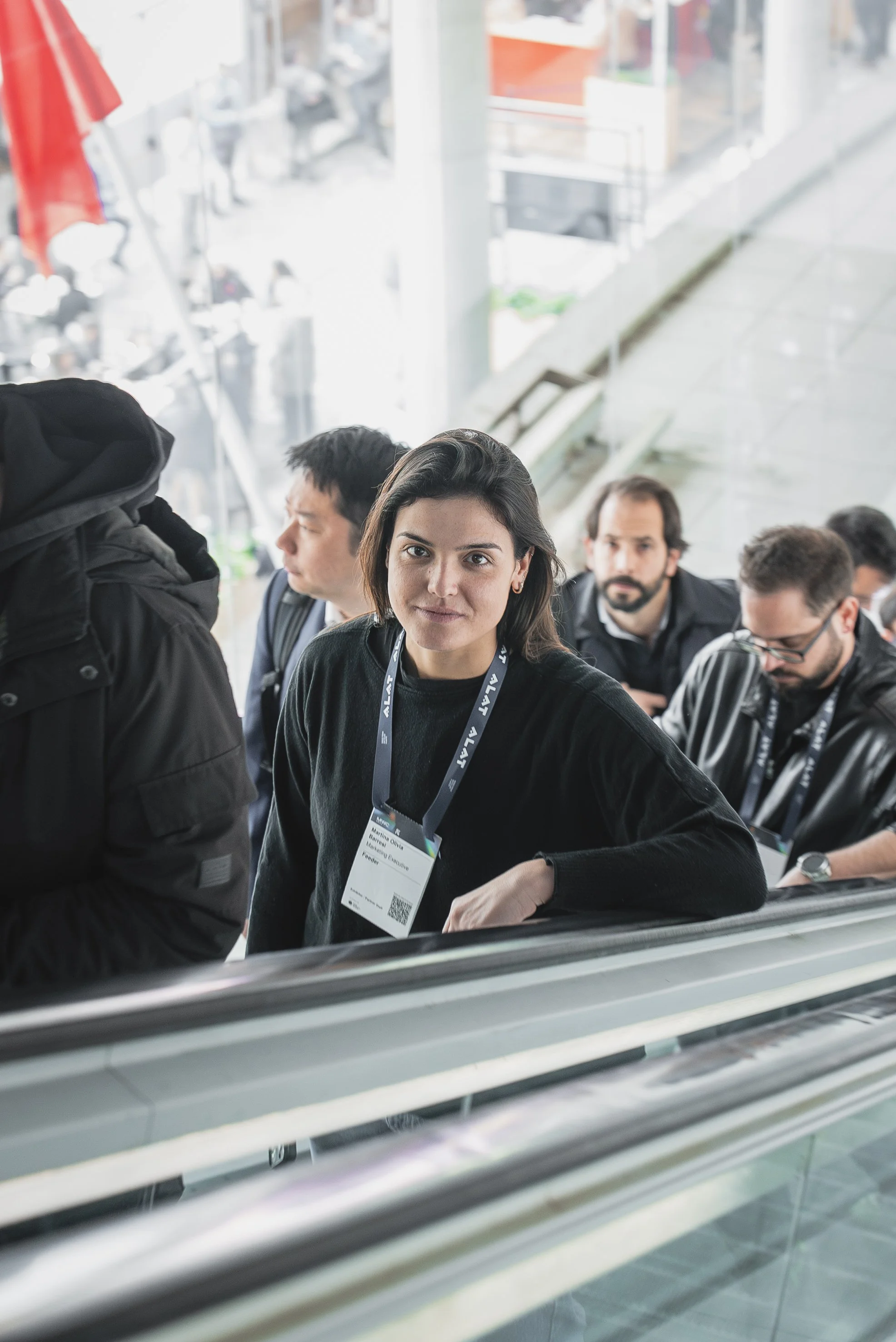 A woman with shoulder-length dark hair and a black sweater, wearing a conference badge, looks at the camera while on an escalator with other people in a modern, glass-walled building.