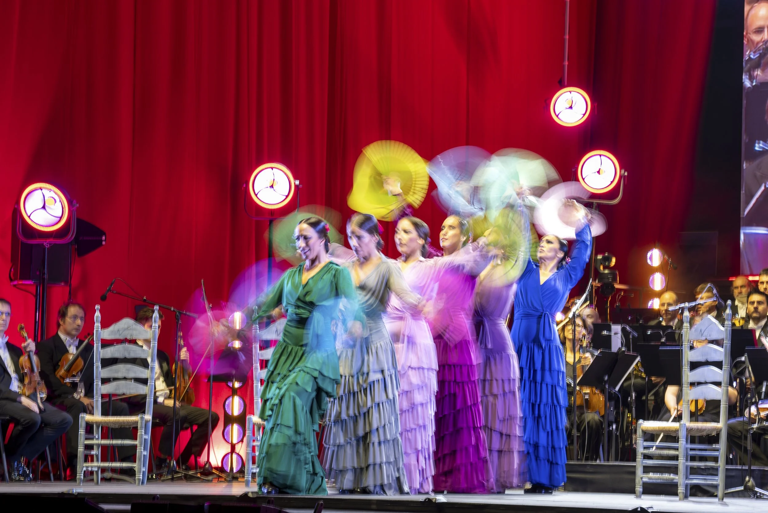 A stage with performers in colorful traditional dresses, an orchestra, and red curtains in the background, with some motion blur indicating movement.