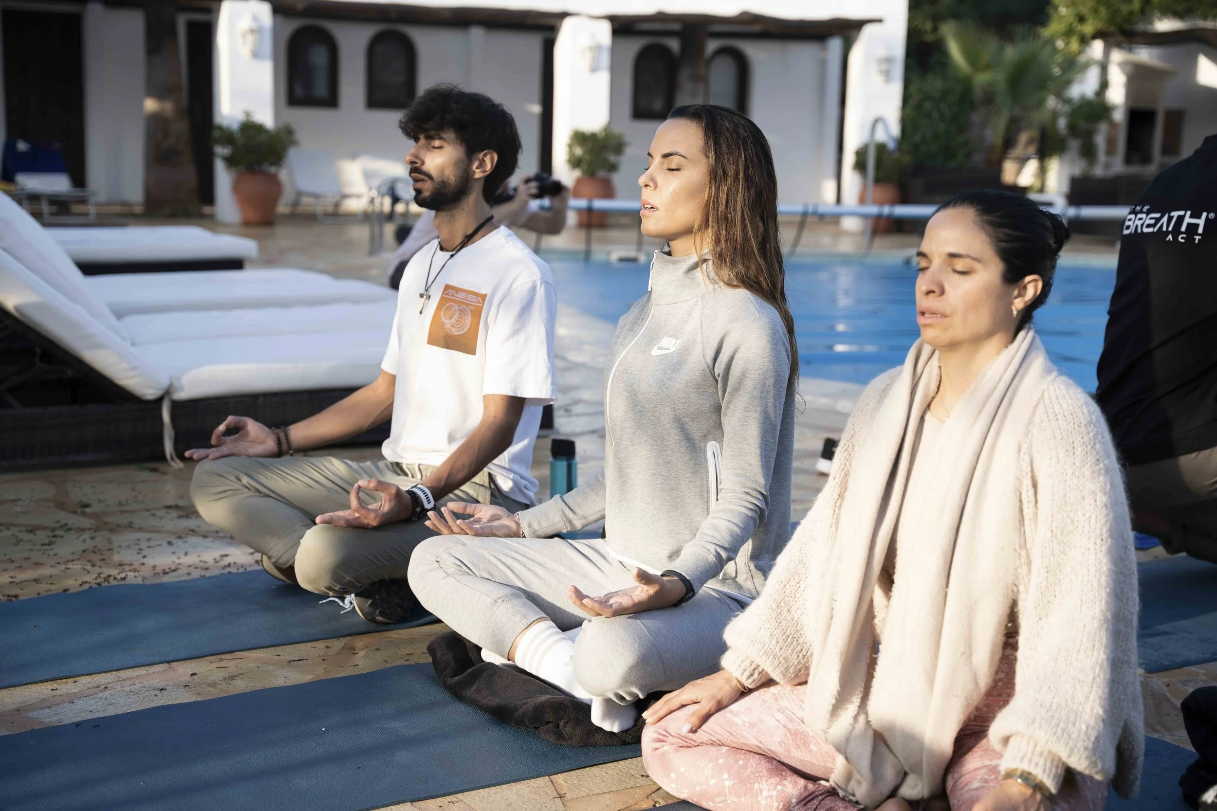 People practicing meditation outdoors by a pool, sitting cross-legged with eyes closed.