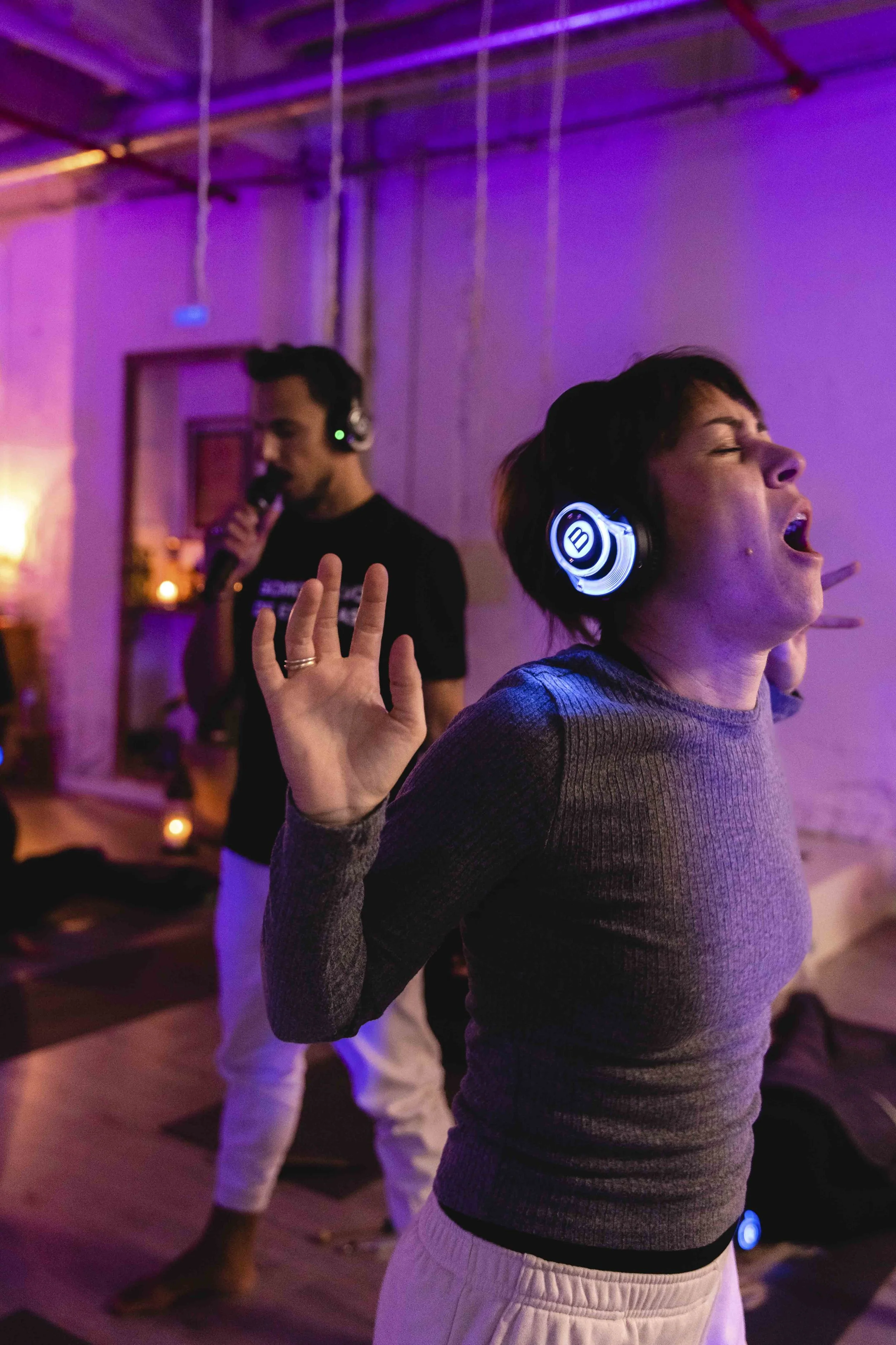 A woman in a gray sweater and white pants has her eyes closed, mouth open, and hands raised as she sings or dances in a room with purple lighting. In the background, a man with headphones and a microphone is singing or speaking, with a hand raised, blurred in the background.