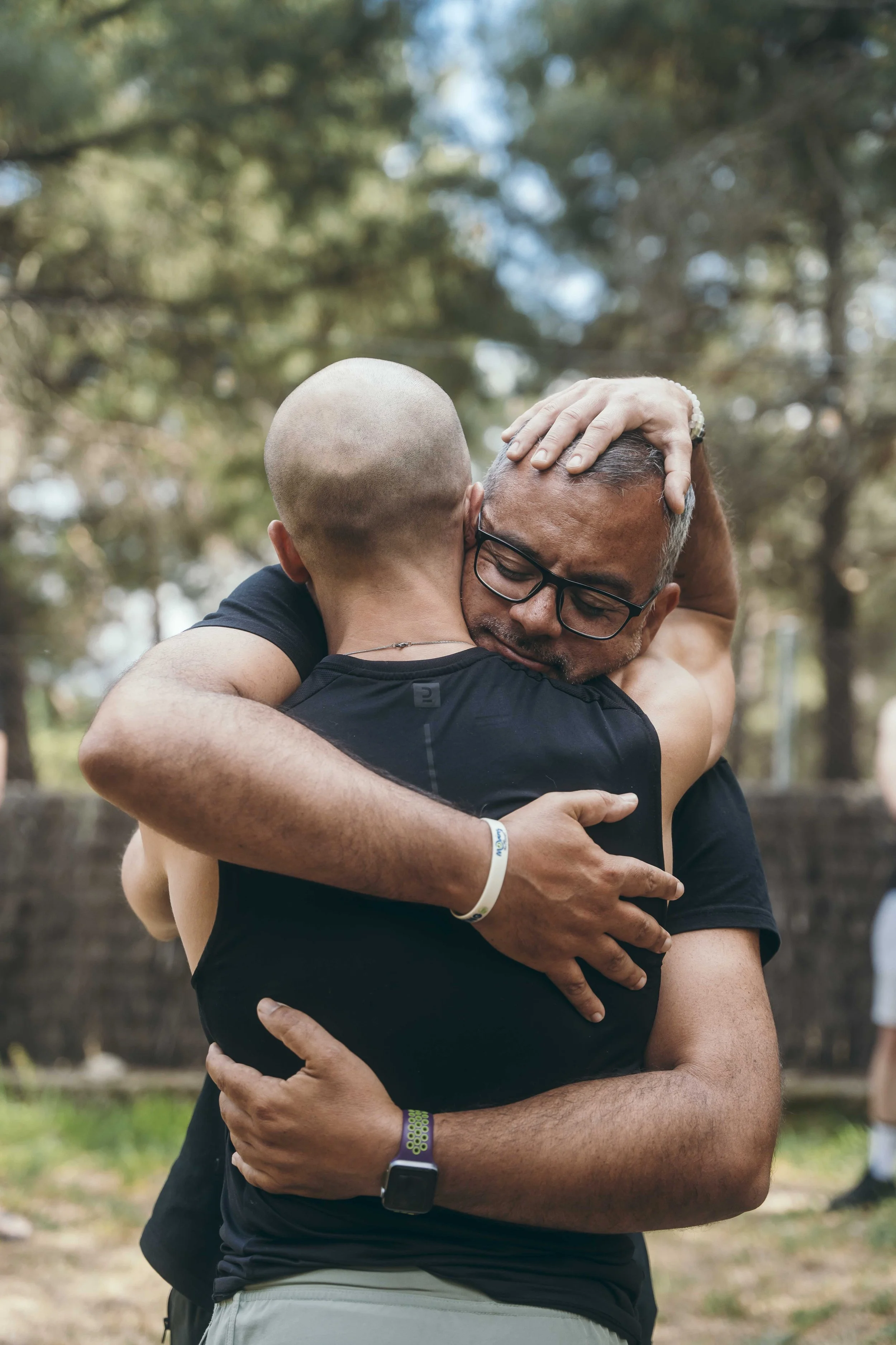Two people hugging outdoors, one with glasses and tattoo, the other bald, in a close, emotional embrace, surrounded by trees.