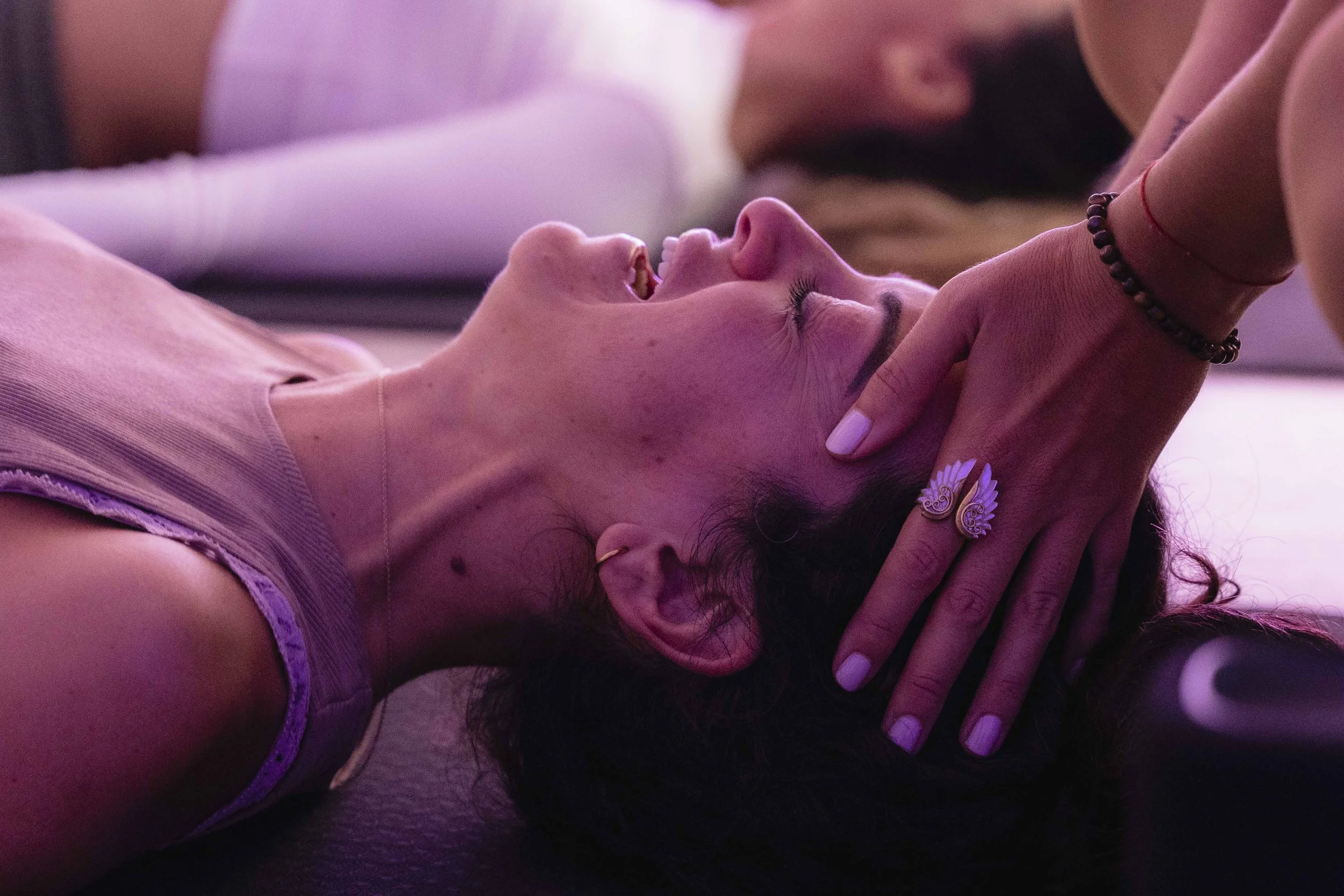 A person lying down with another person holding their face, close-up shot with pink and purple lighting.