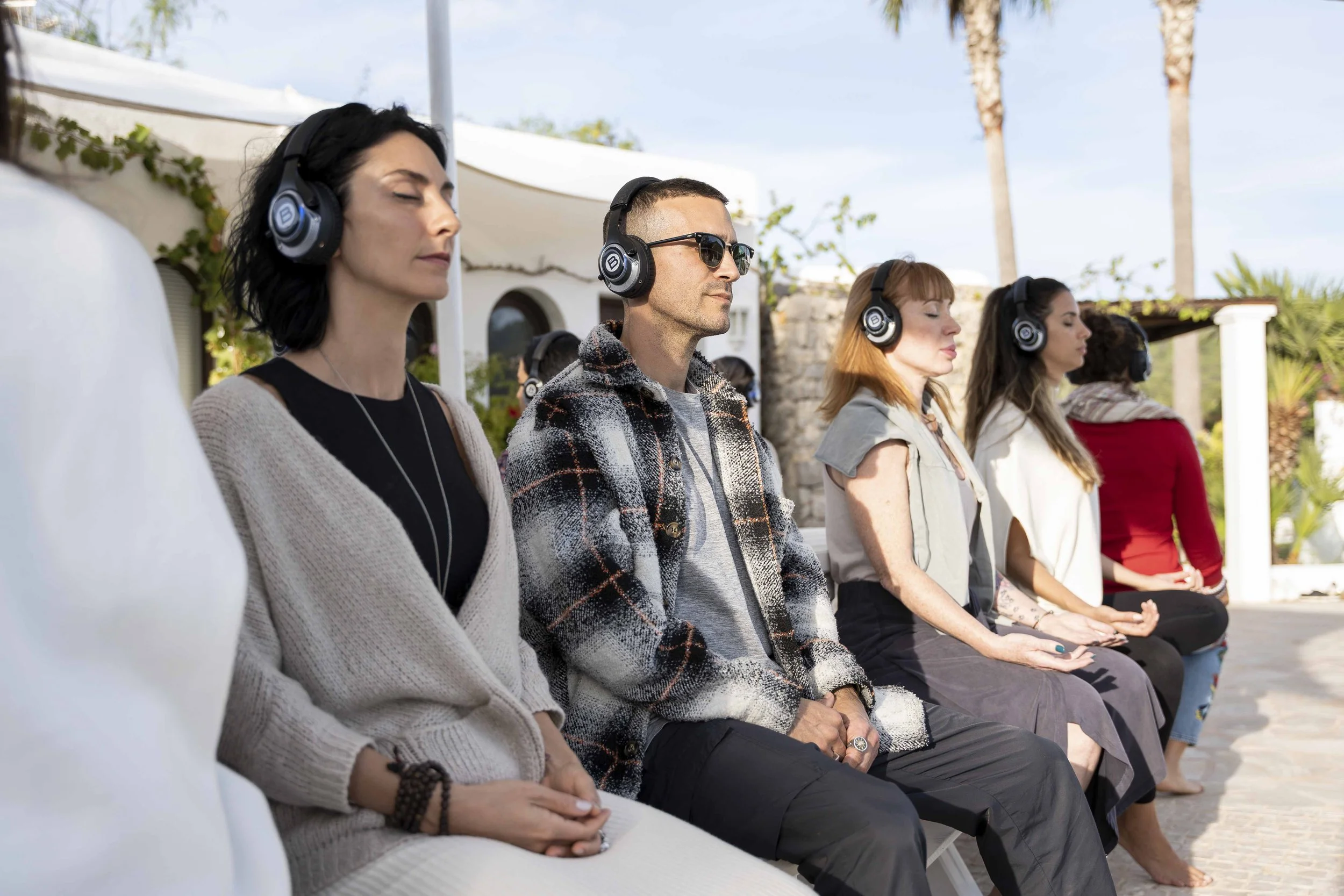 A diverse group of people sitting outdoors, meditating with eyes closed and relaxed expressions, under a white canopy with palm trees in the background.