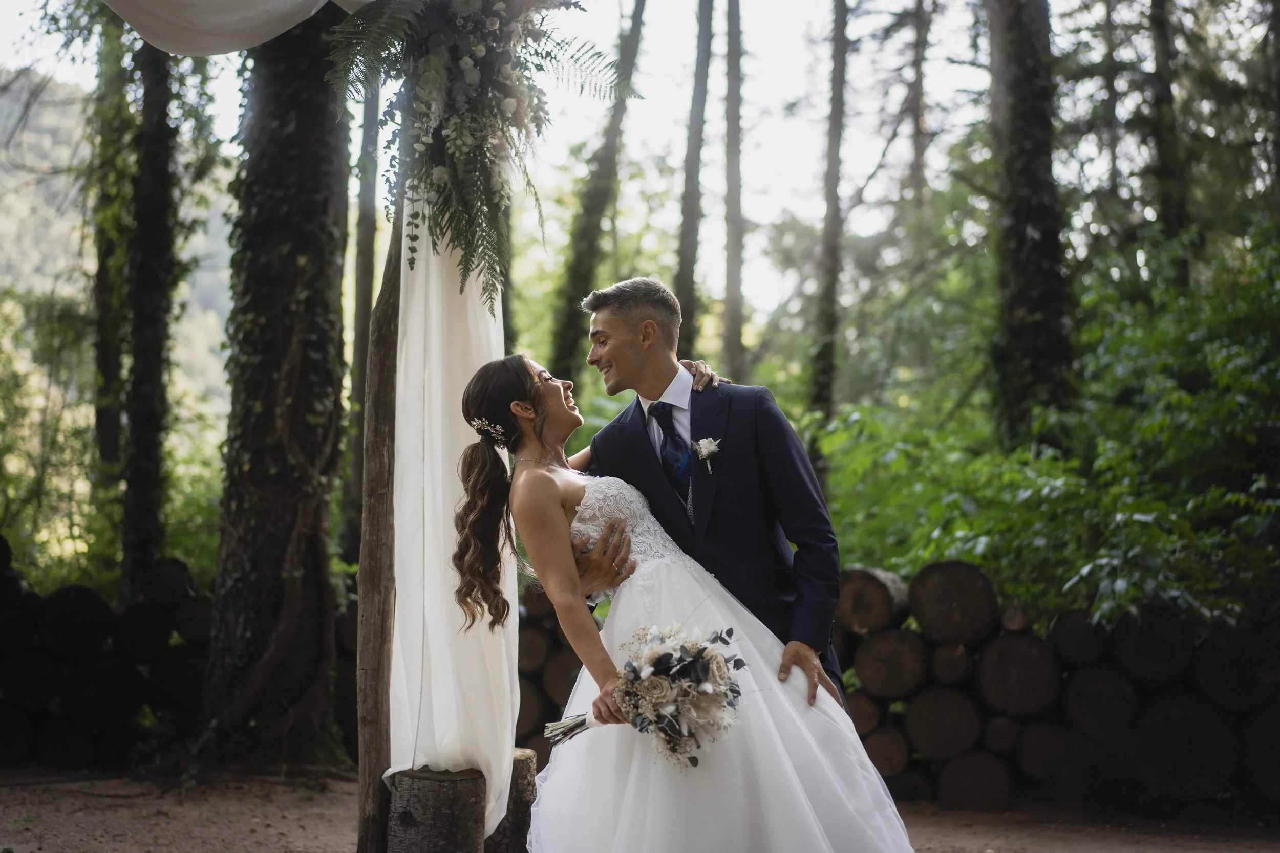 A bride and groom are dancing outdoors during their wedding ceremony in a forest setting, with the bride holding a bouquet and the groom smiling.