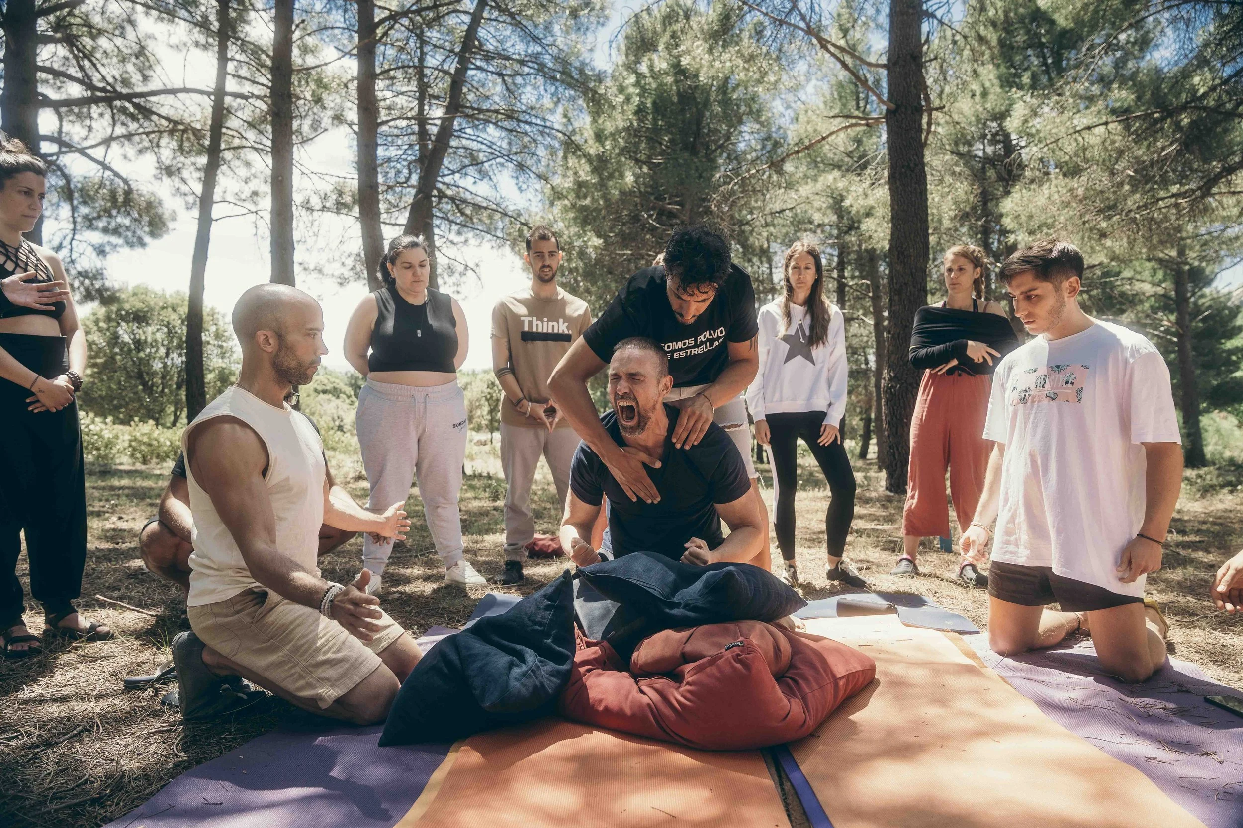 A group of people gathered outdoors in a forested area, participating in a demonstration or rally. Two men are kneeling on the ground in the center, with one of them holding a microphone and the other hand on his chest, appearing to be speaking or singing. The group is diverse and standing around them, some looking serious and attentive.